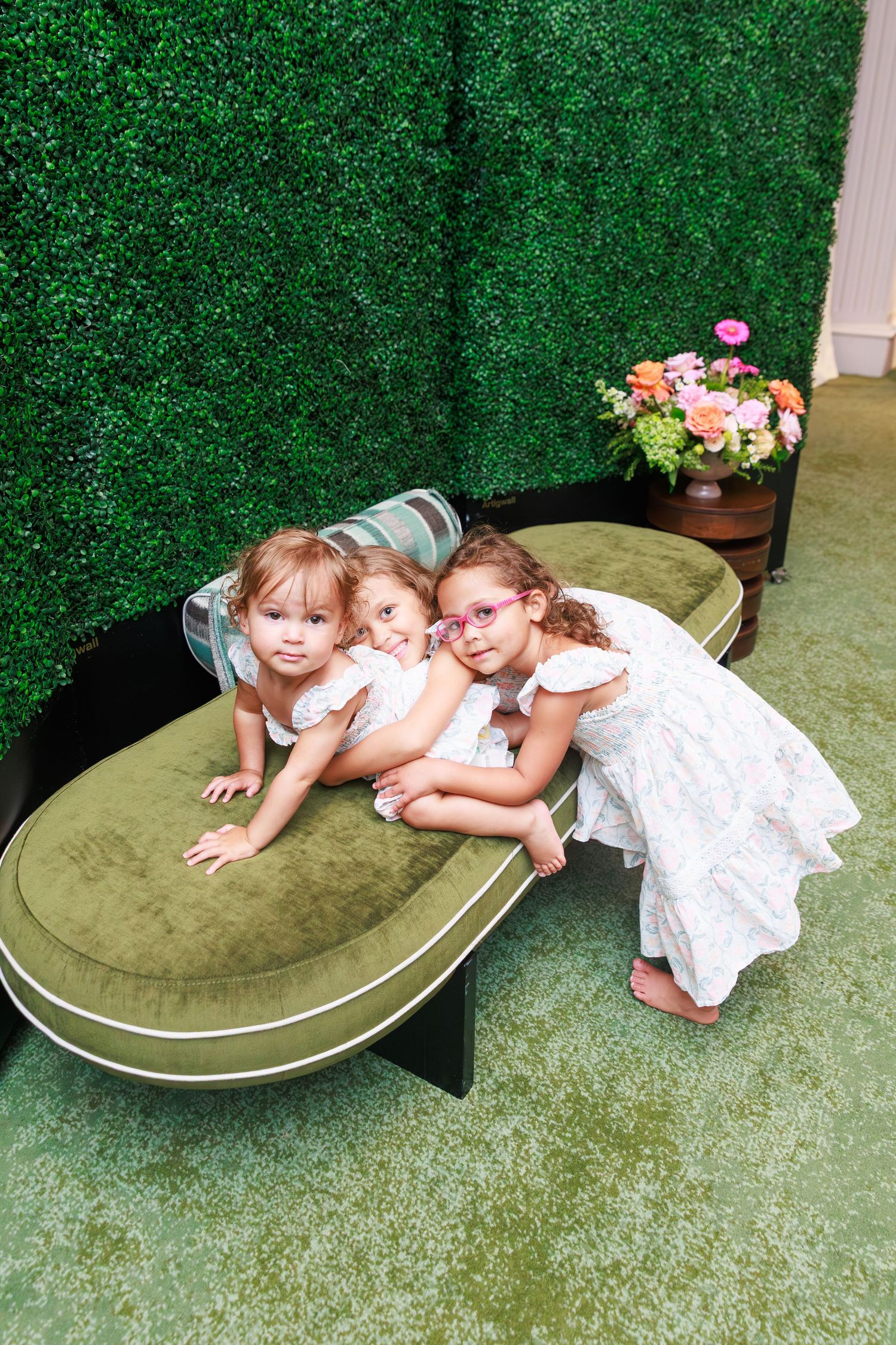 Three young girls in white dresses pose together on a green velvet bench against a lush hedge wall backdrop.