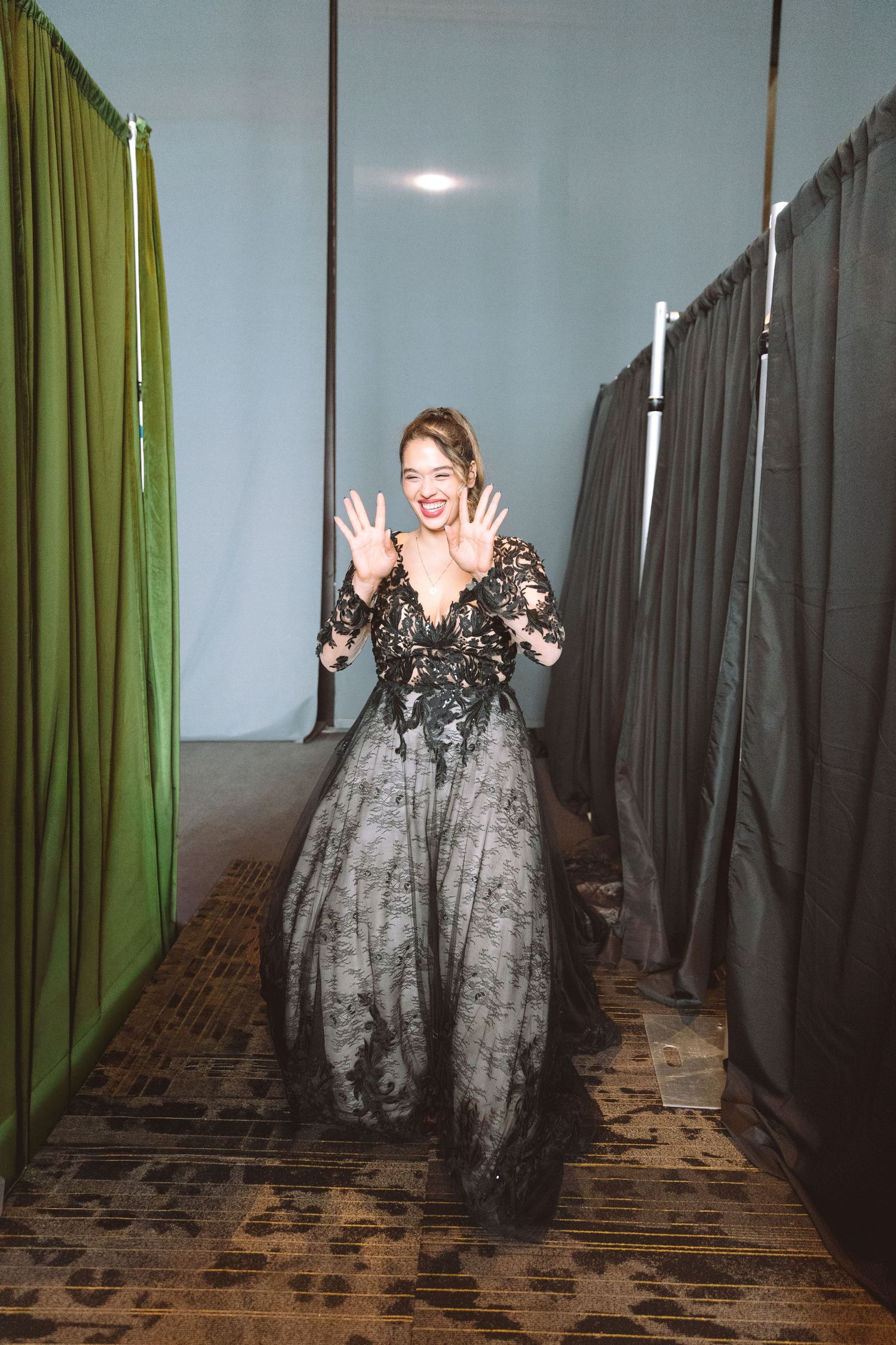 Woman in elegant black lace ballgown posing with hands raised in a dressing room area.