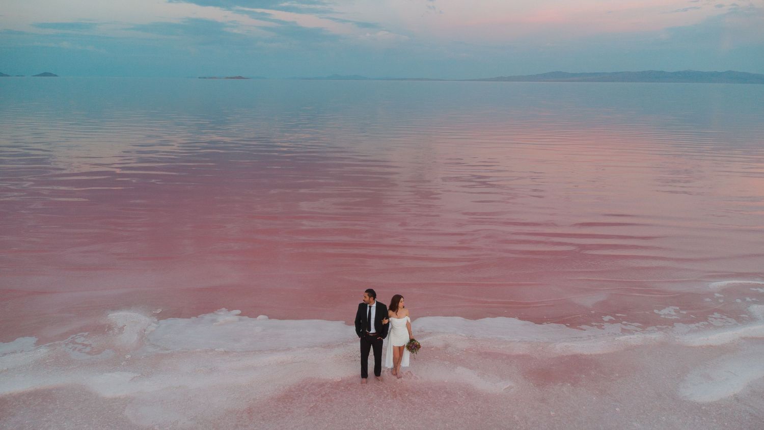 Couple in wedding attire standing on pink salt lake shore at sunset with vibrant pink water and sky.
