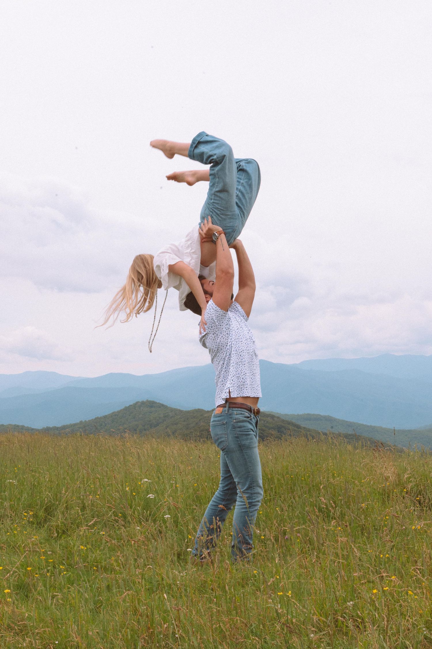 Man lifting woman high above his head in a grassy mountain meadow under cloudy skies.