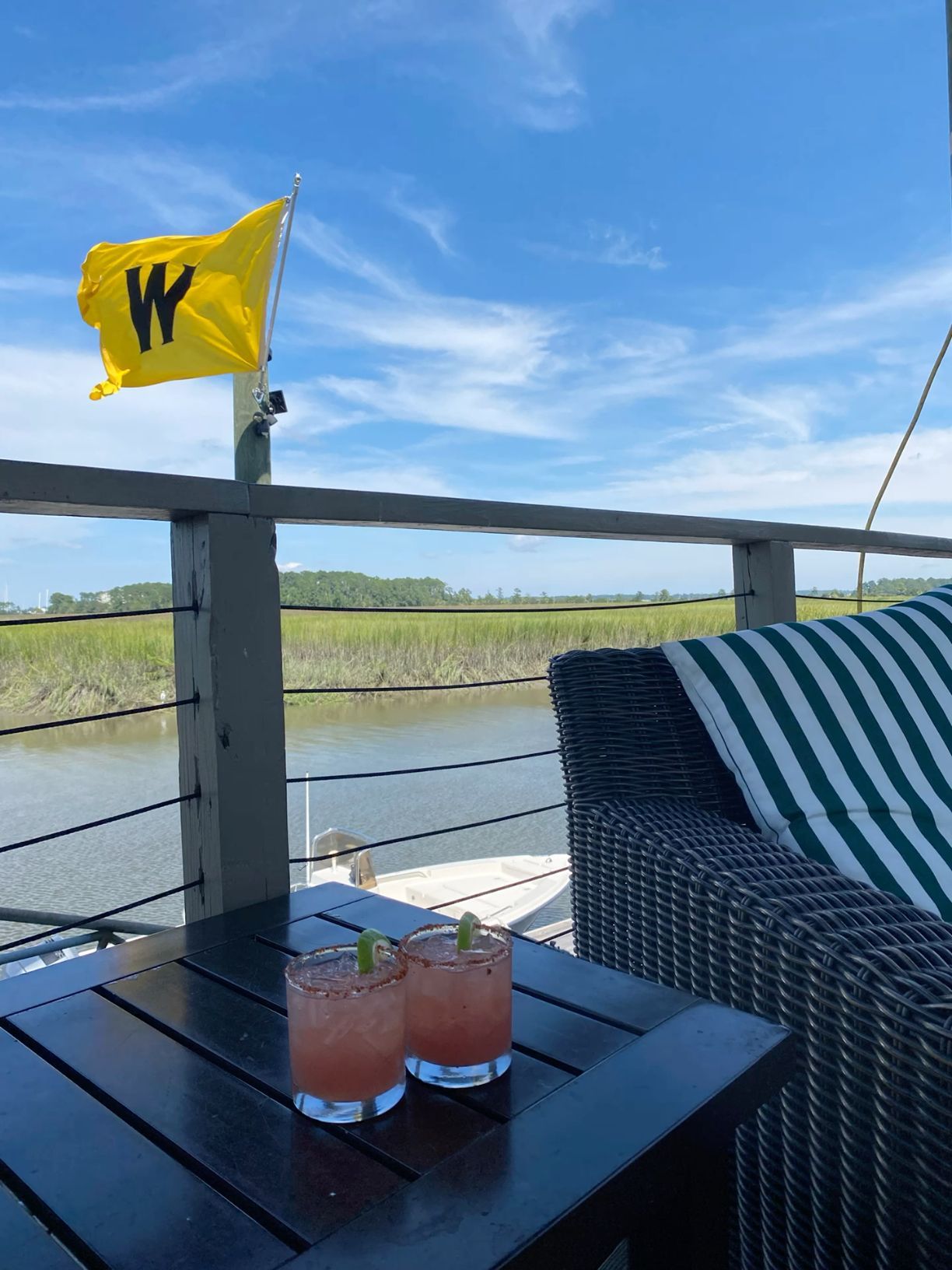 Two pink cocktails on deck table overlooking marsh with yellow W flag under blue sky.