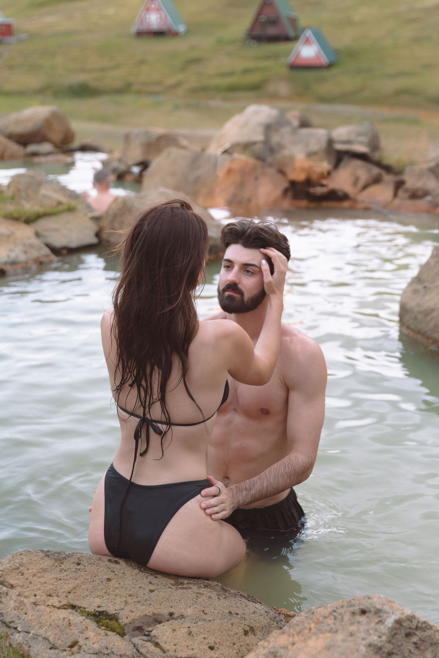 Woman in black bikini touching mans hair while sitting together in hot spring.