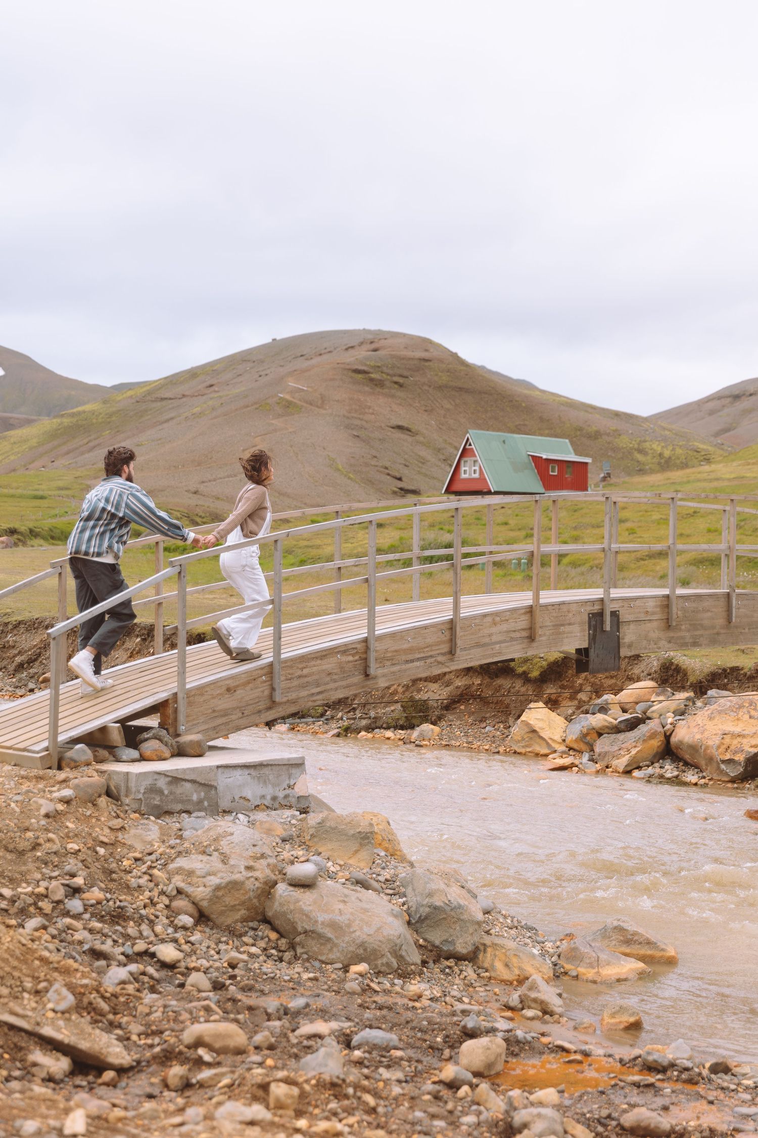 Couple holding hands crossing a wooden bridge near a red A-frame cabin in an Icelandic highland landscape.