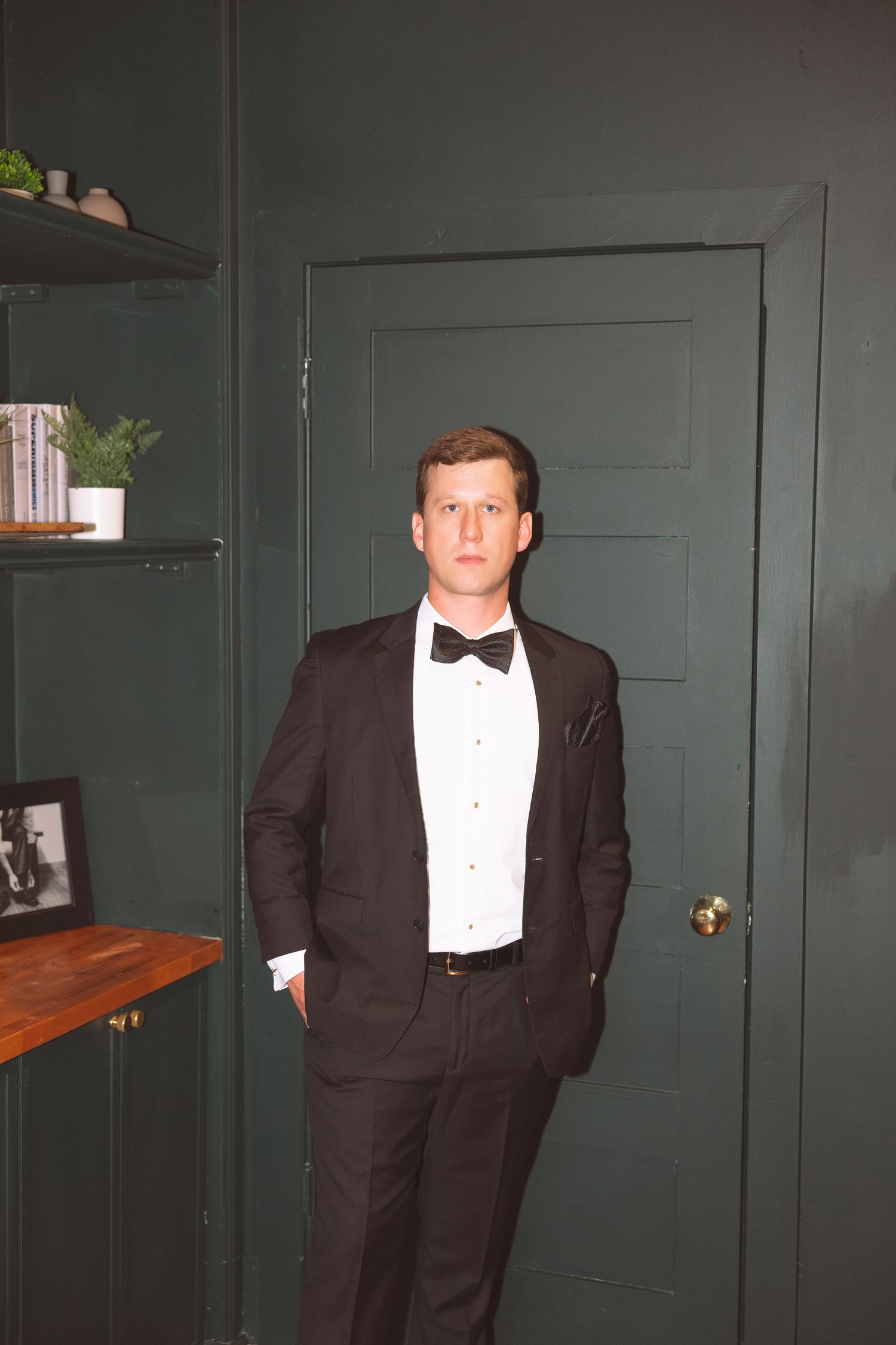 Groom in classic black tuxedo with bow tie poses confidently in front of dark green door.