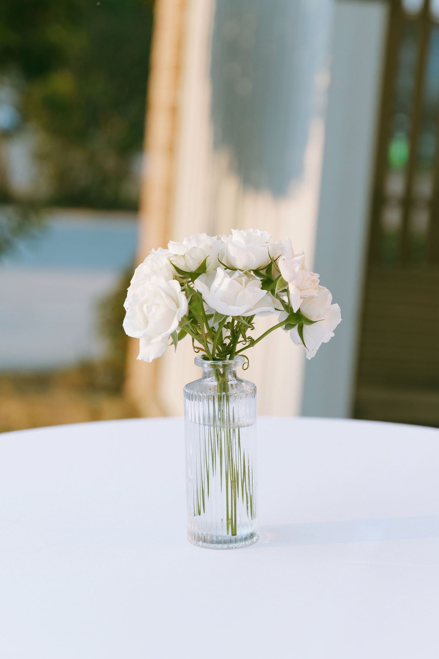 Small glass ribbed vase with white roses centerpiece on white tablecloth at an outdoor wedding reception.