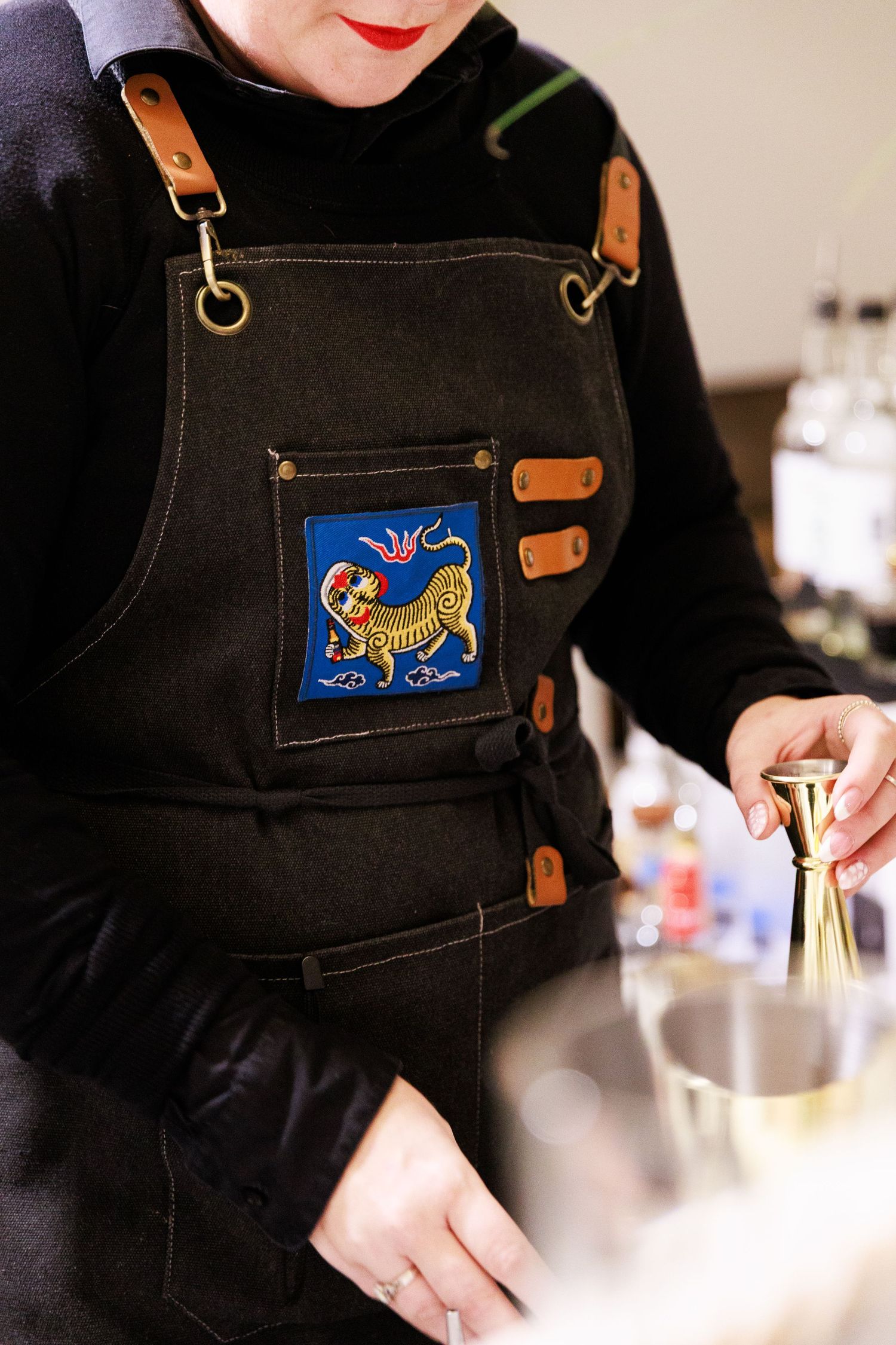 Bartender in black apron with tiger patch uses gold jigger to mix cocktails at a bar.