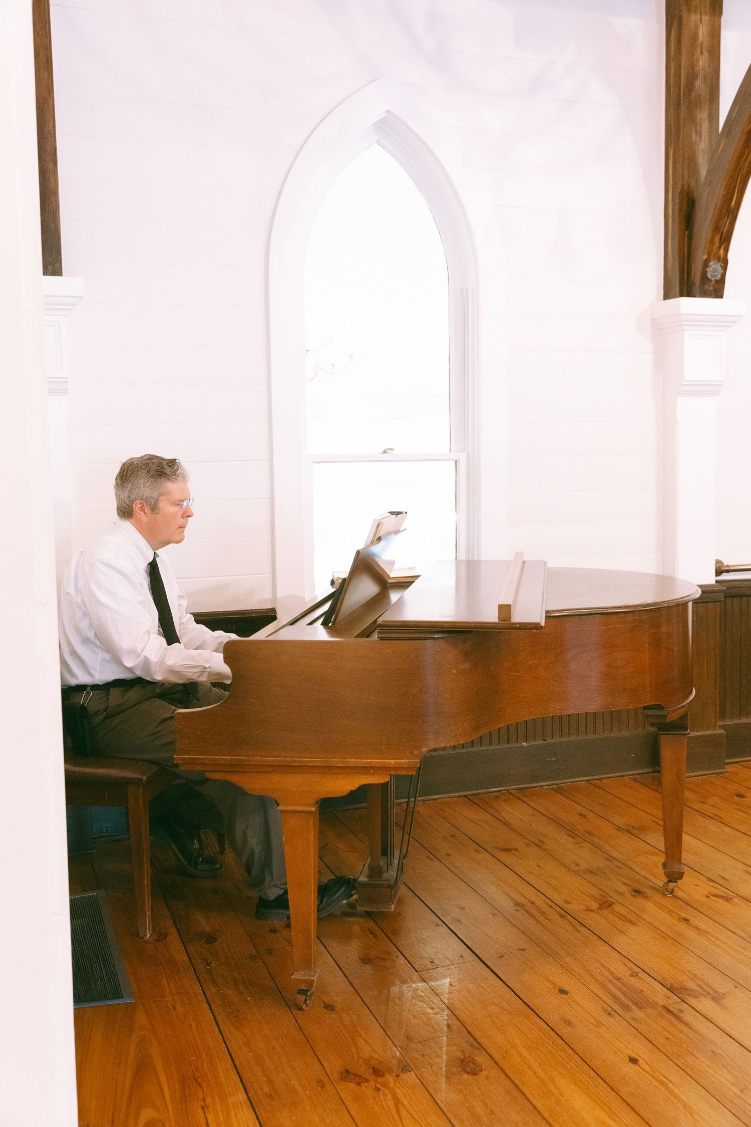 Elderly man in dress shirt and tie playing a grand piano in a white church with arched windows and hardwood floors.