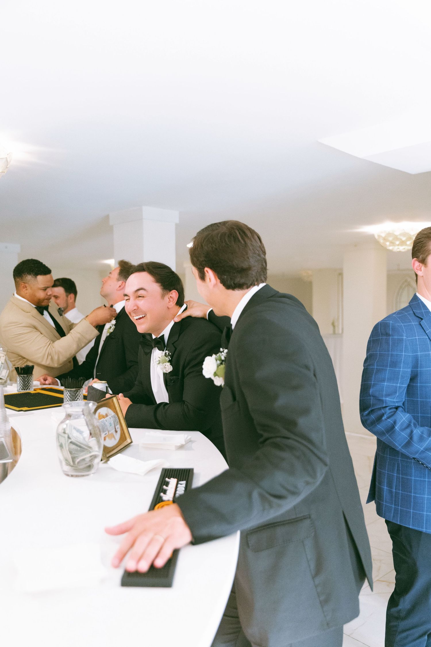 Groom laughing with guest at elegant indoor wedding reception near white tablecloth bar.
