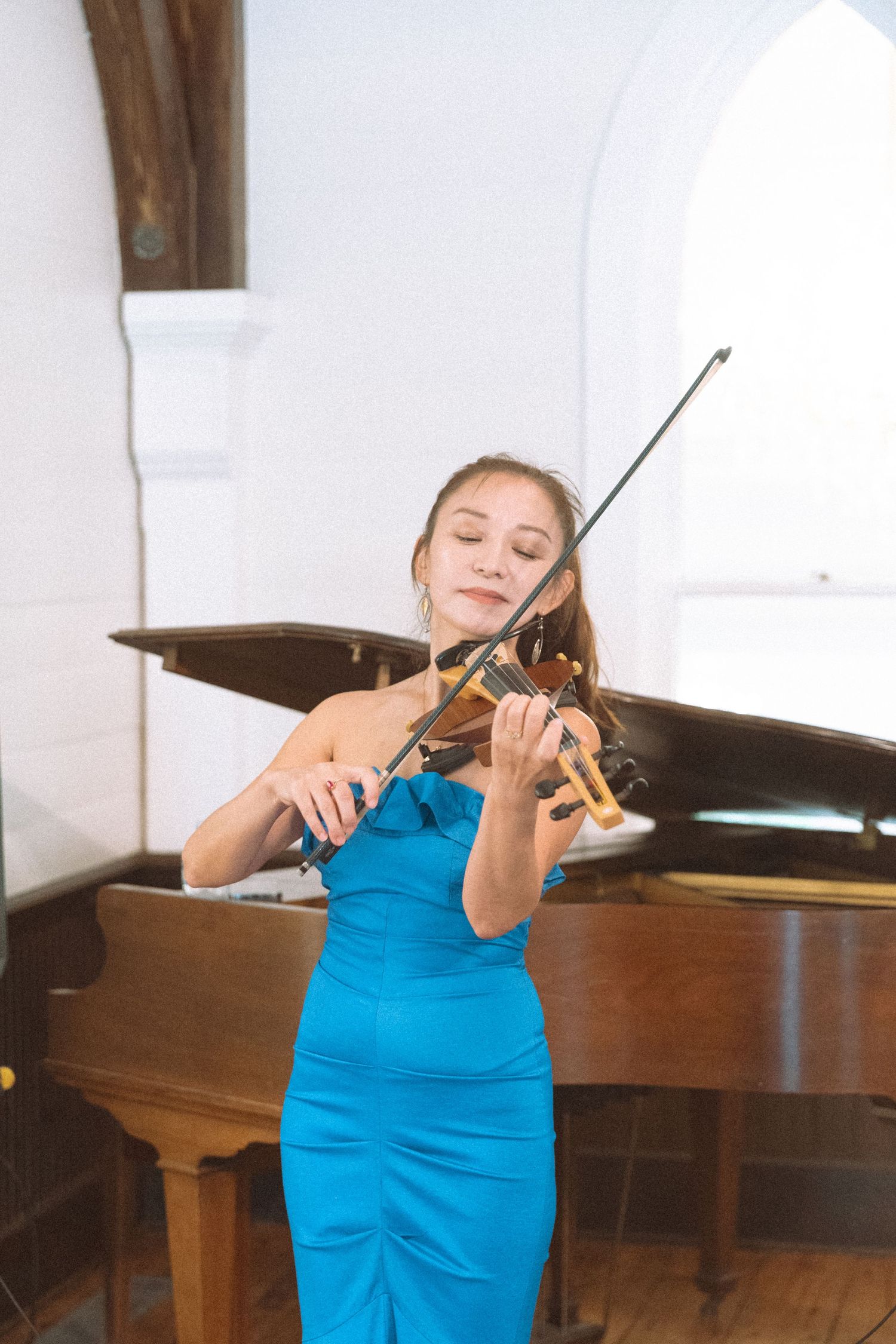 Young woman in blue dress playing violin in front of a grand piano in a bright white room.