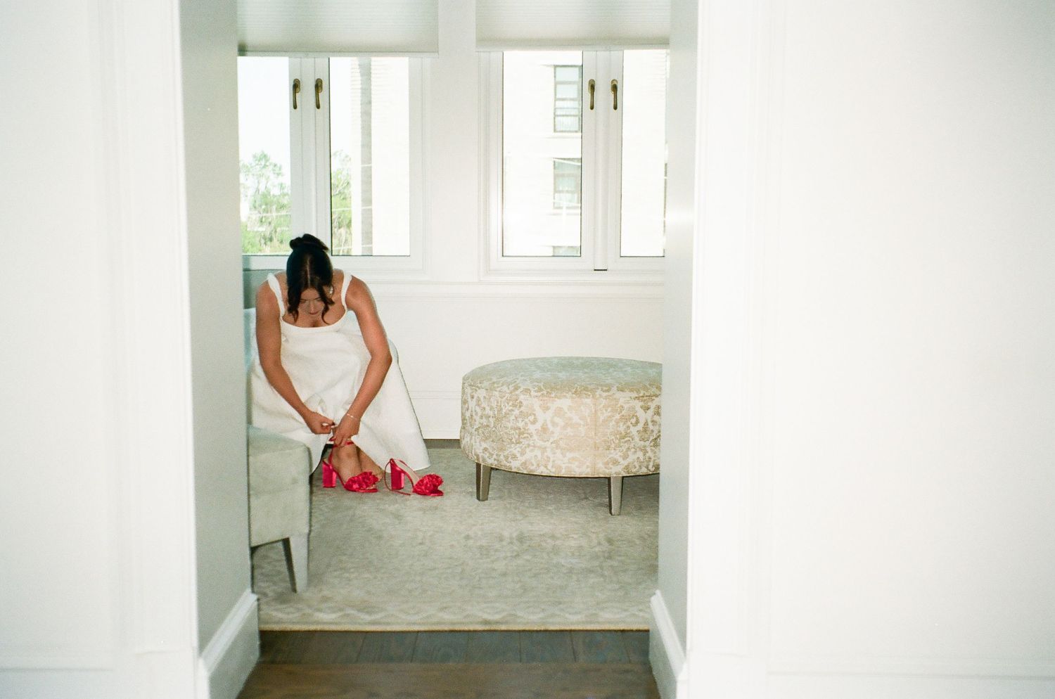 Woman in white dress putting on red heels in a bright, elegant dressing room with an upholstered ottoman.