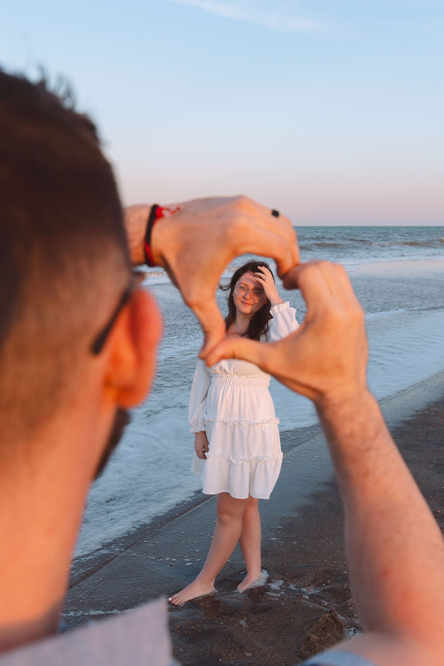 Man framing woman in white dress on beach at sunset with ocean waves in background.