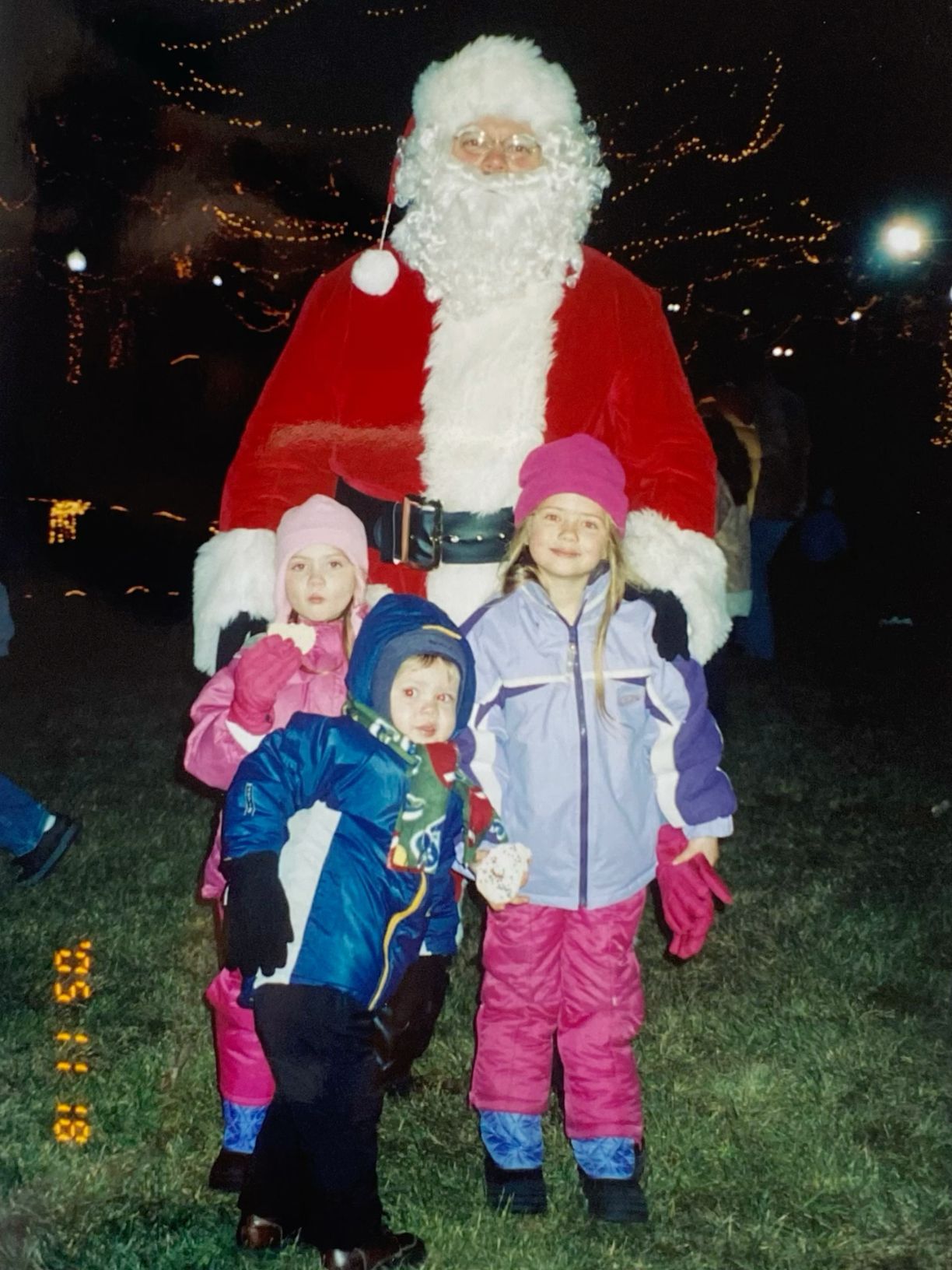 Three children pose with Santa Claus outdoors at a Christmas event, wearing winter coats with holiday lights in background.