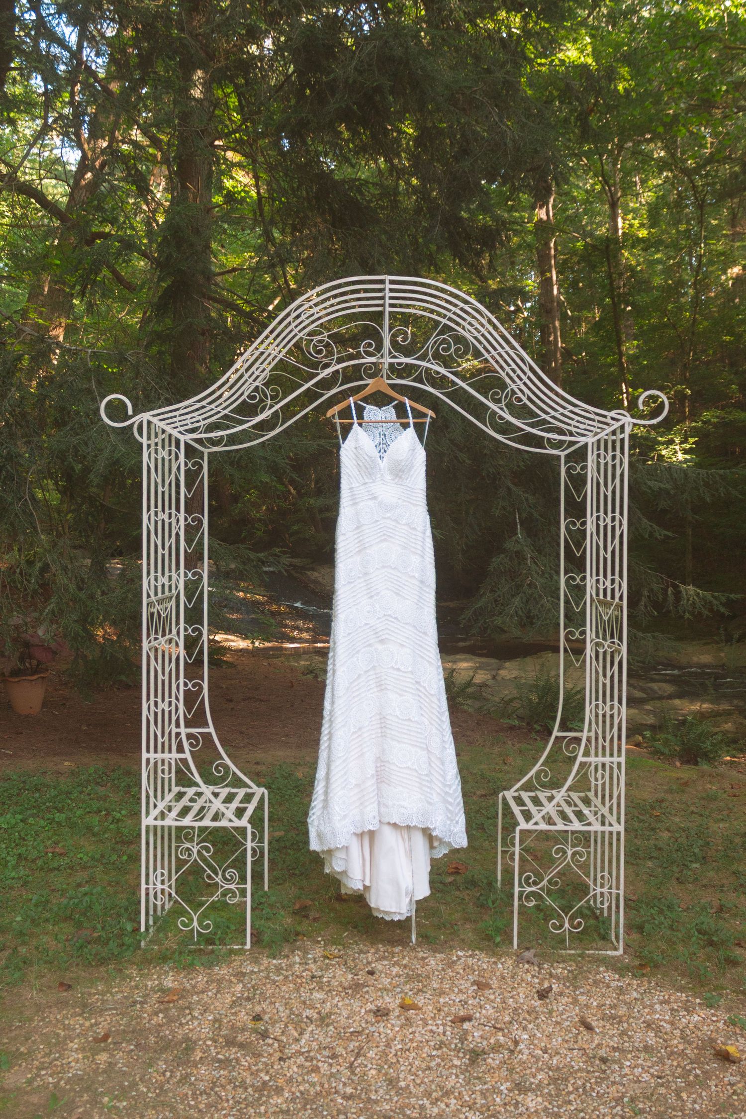 White lace wedding dress displayed on ornate wrought iron garden arch in wooded outdoor setting.