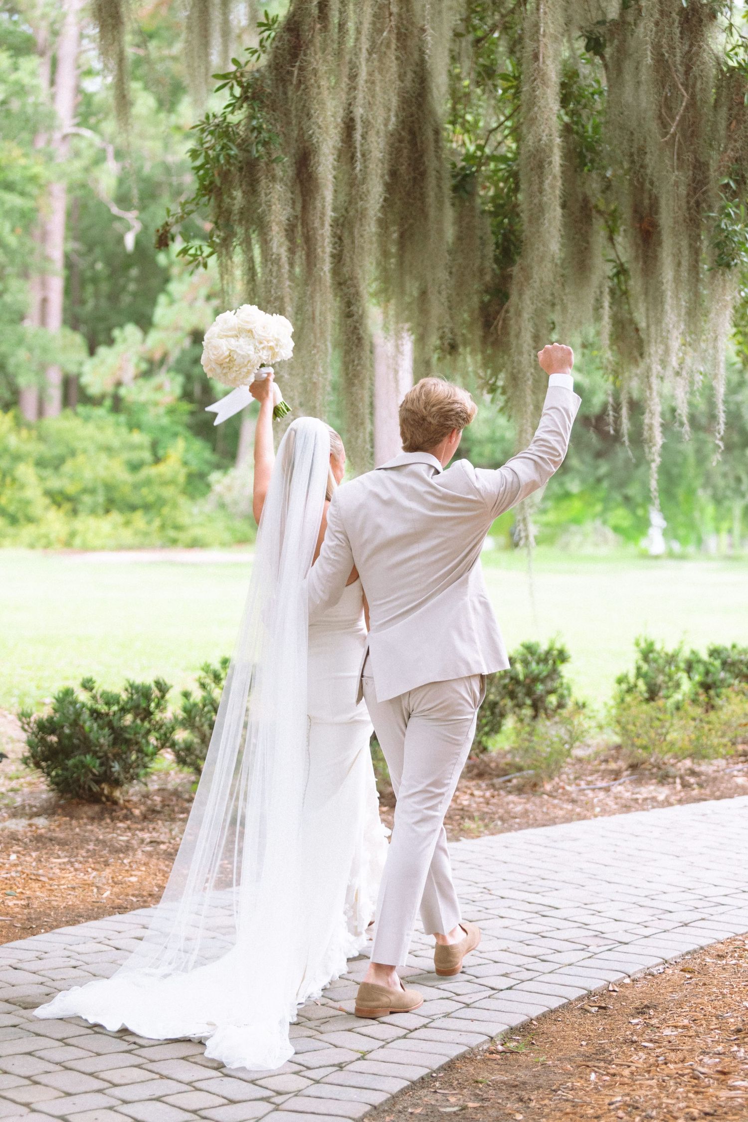 Bride and groom celebrating walking away under Spanish moss trees, arms raised in joy.