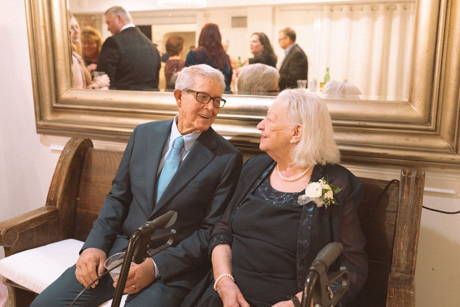 Elderly couple sitting together at wedding reception beside ornate gold-framed mirror.