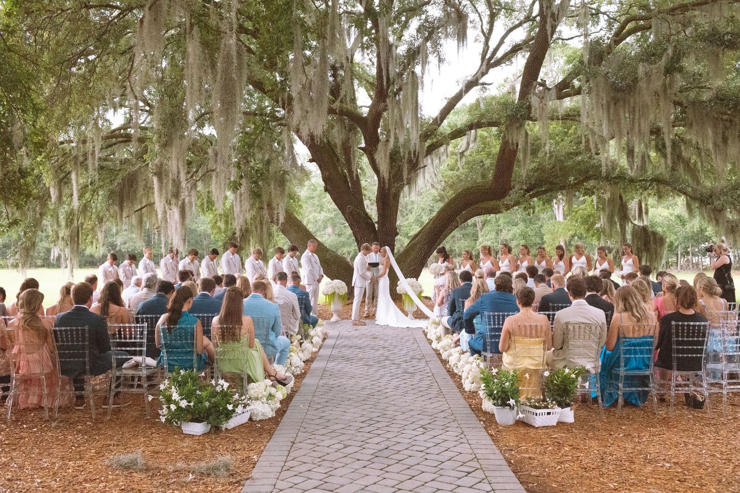 Outdoor wedding ceremony under sprawling oak trees with Spanish moss, guests seated along cobblestone aisle in lush garden setting.