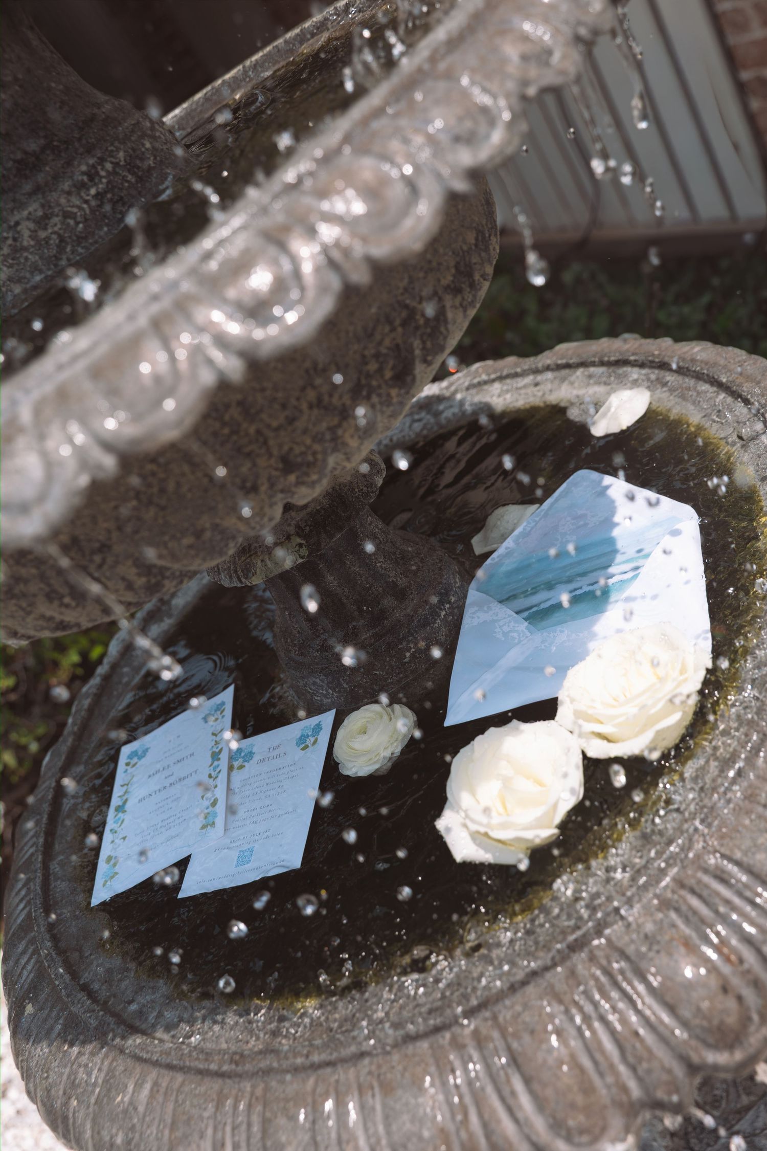 Wedding invitations and white roses rest in a stone water fountain.