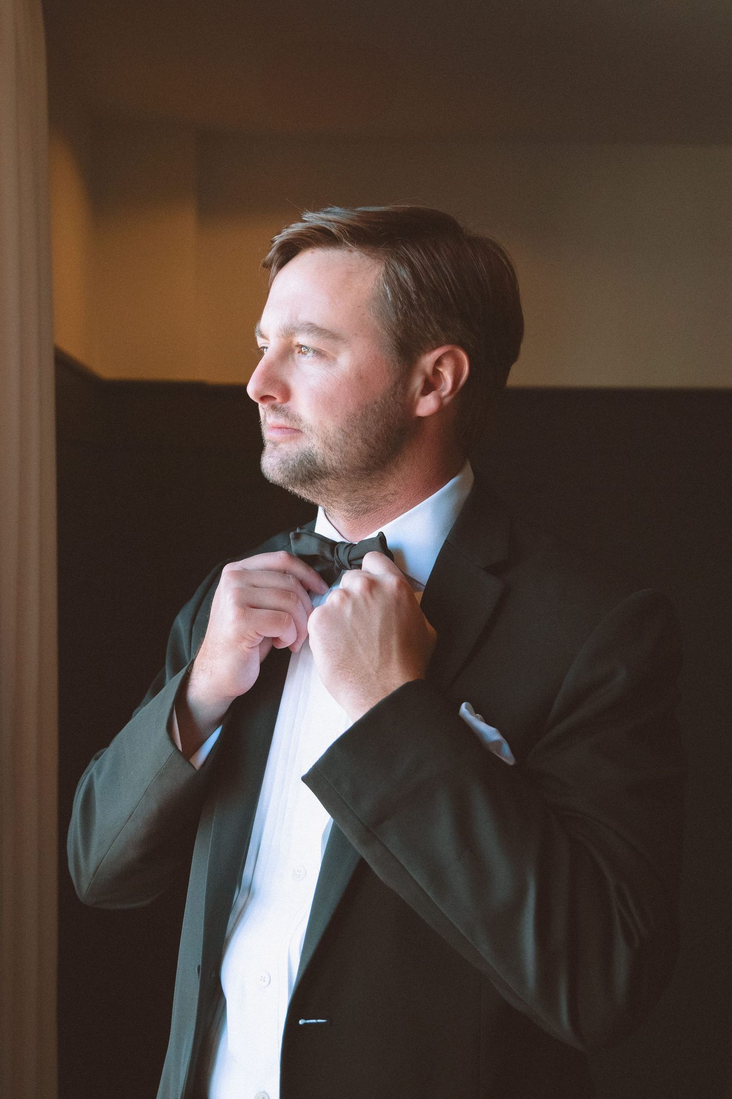 Dapper groom adjusts his black bow tie while getting ready in warm indoor lighting.