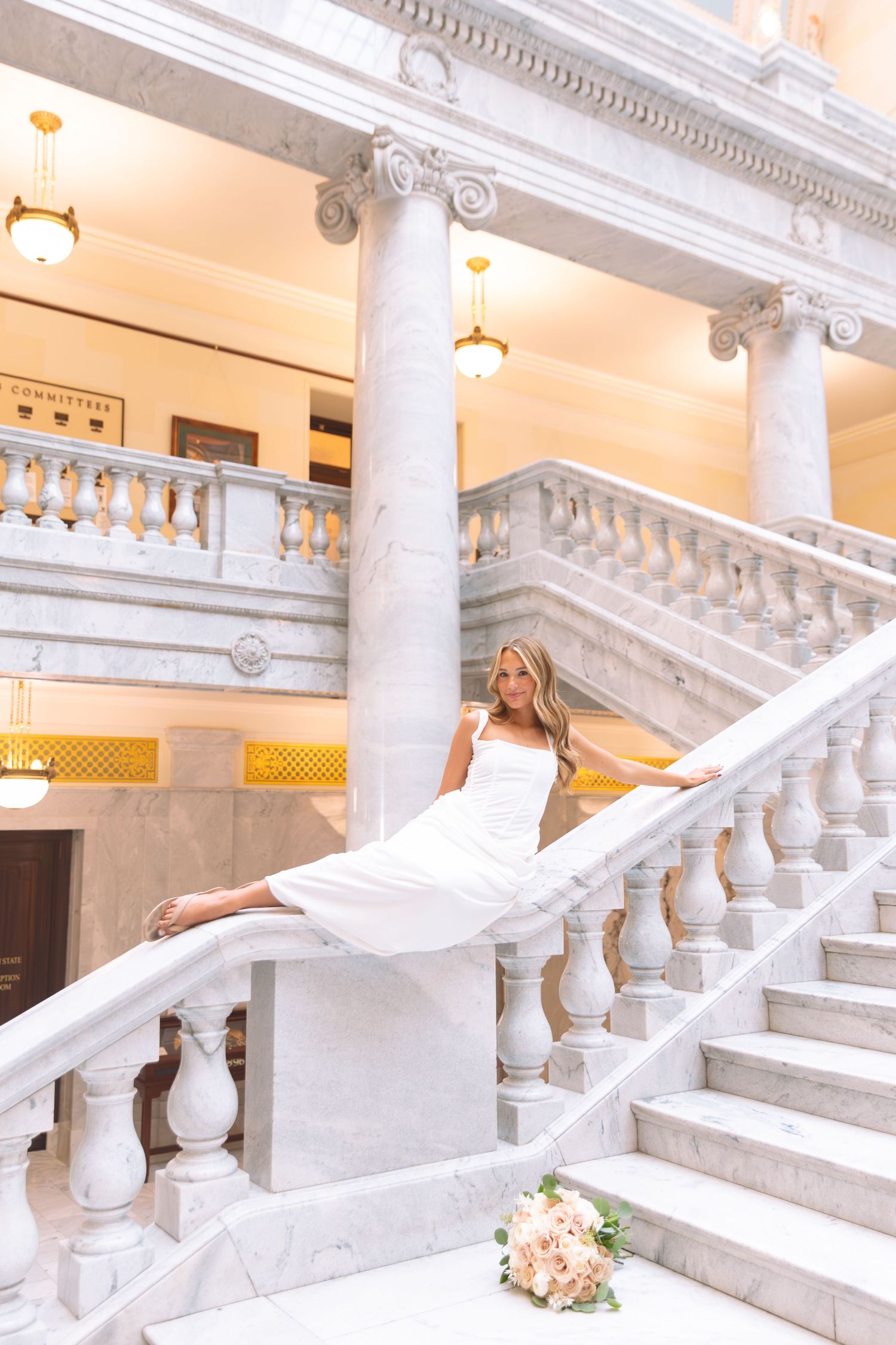 Elegant bride in white dress posing on grand marble staircase with classical columns and bouquet.