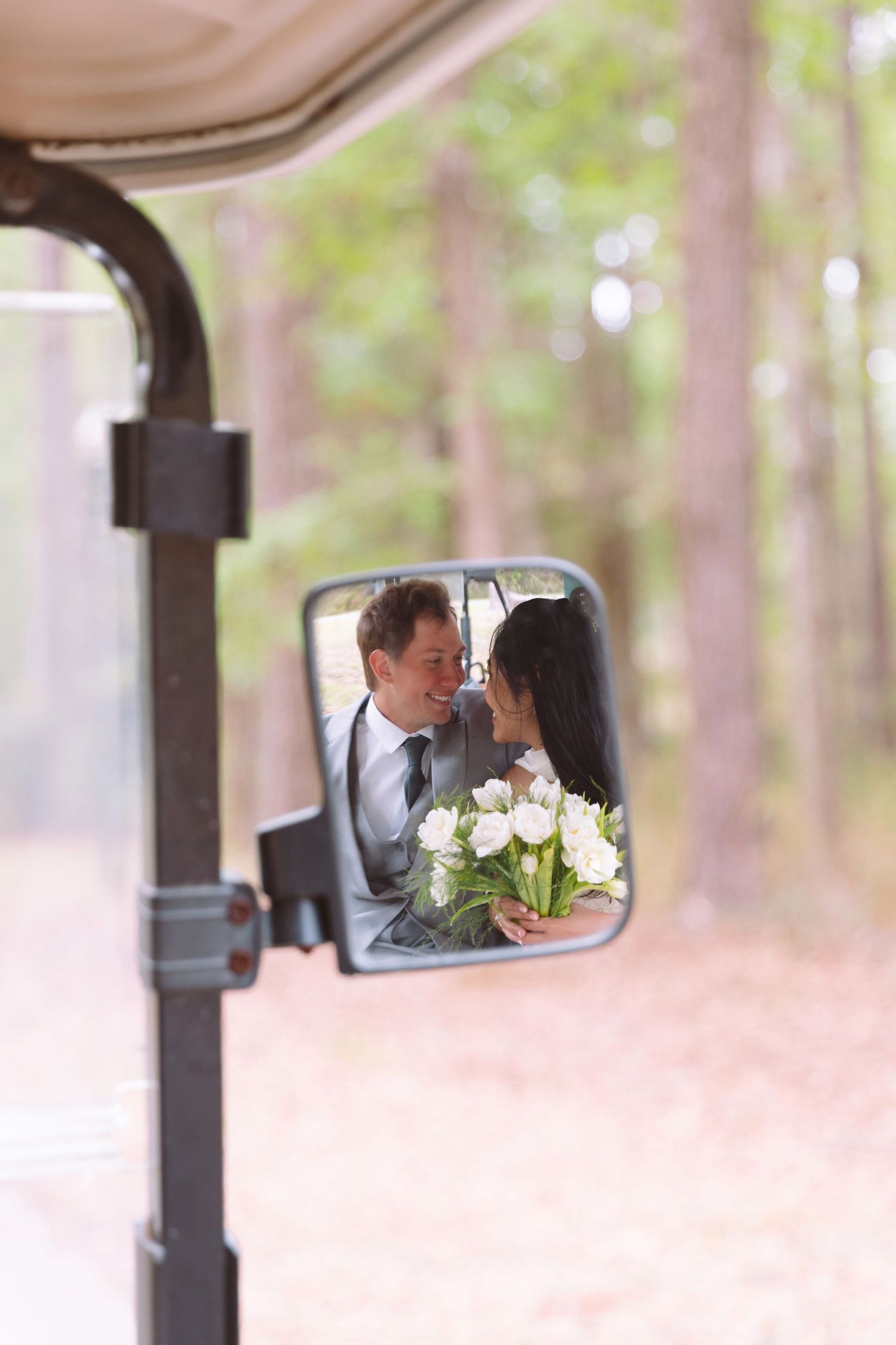 Bride and groom reflected in a golf cart mirror, surrounded by pine trees, bride holding white tulip bouquet.