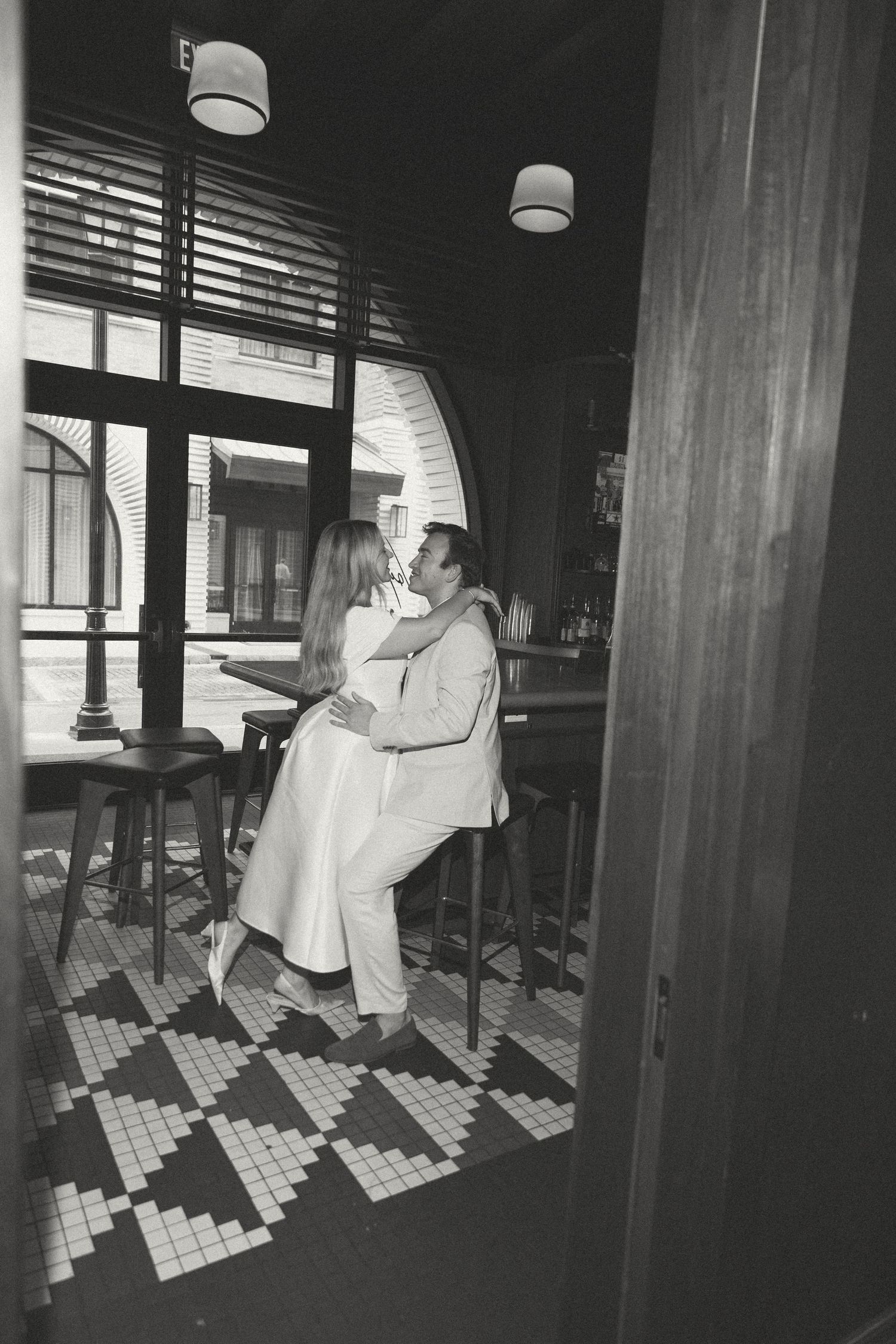 Black and white photo of a couple embracing romantically inside a stylish bar with mosaic tile floors.