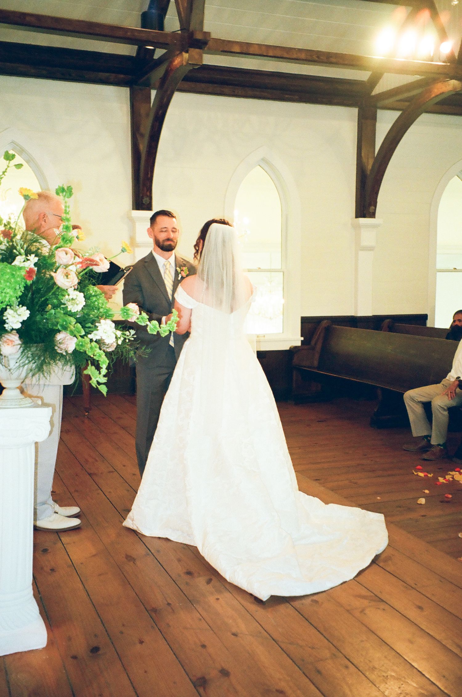 Bride and groom exchange vows in a charming white chapel with wooden floors, arched windows, and lush floral arrangements.