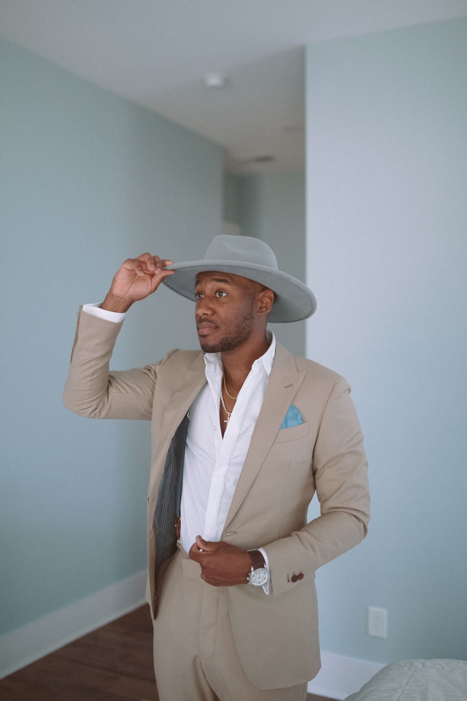 Elegant man in beige suit touching brim of gray wide-brim hat, wearing blue pocket square and gold necklace.