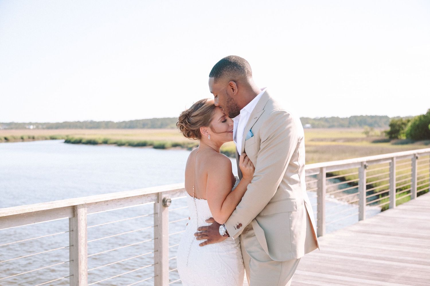 An interracial newlywed couple shares a tender moment on a waterfront dock in bright natural light.