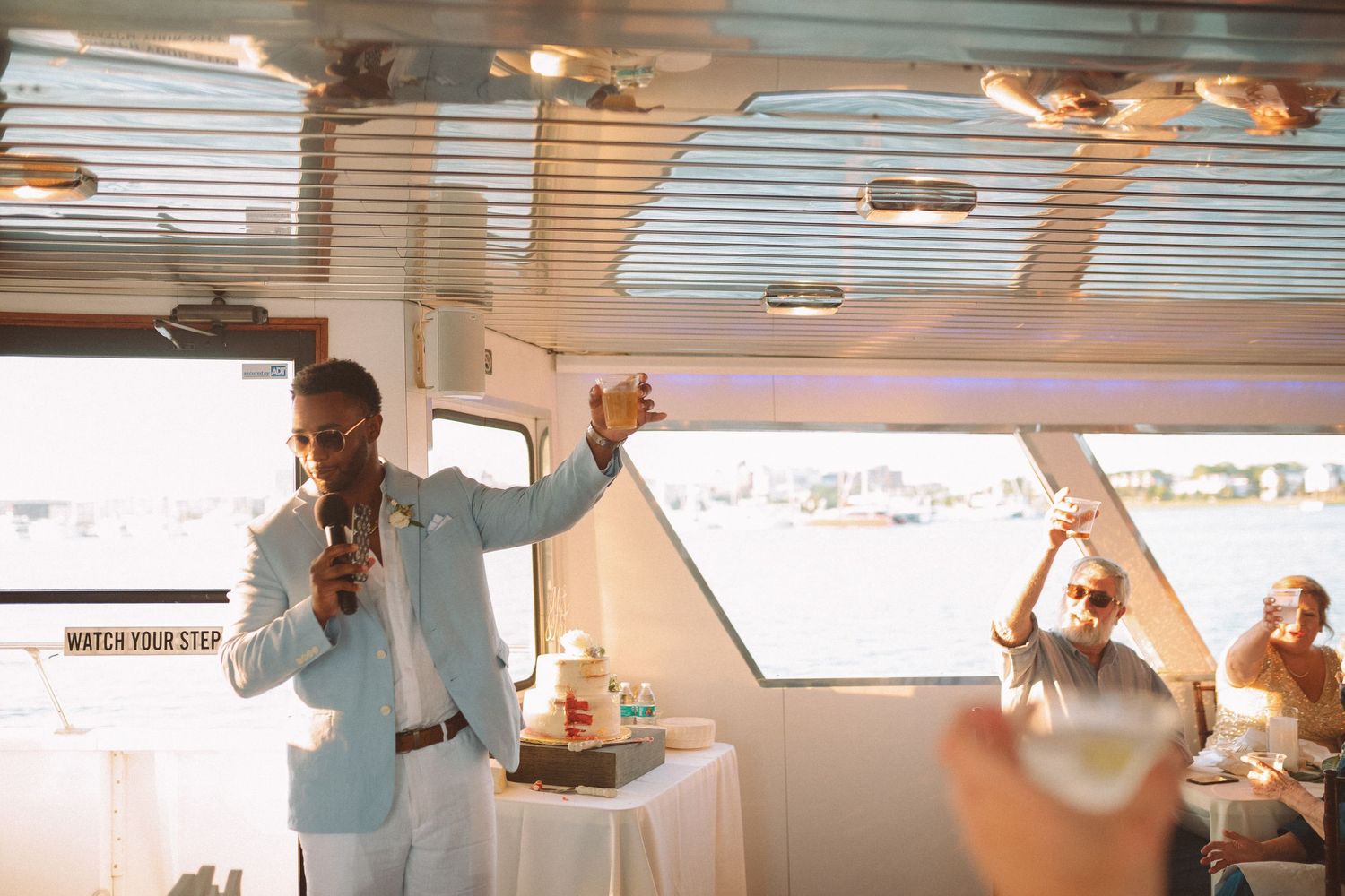 Guest gives energetic speech with microphone while crowd cheers at boat wedding reception.