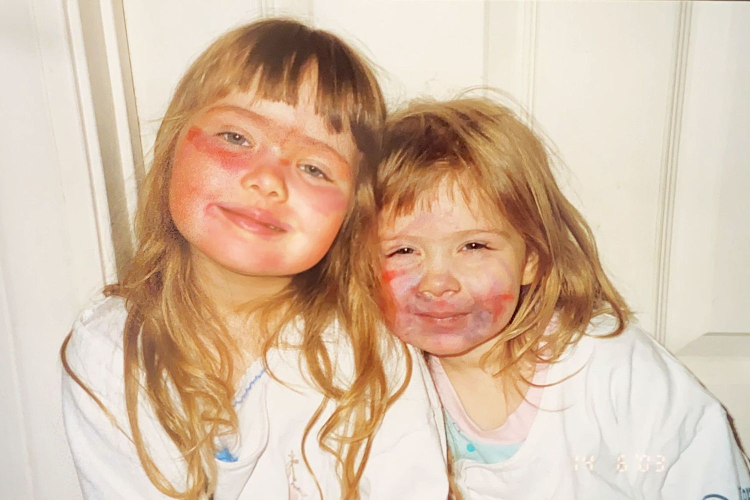Two blonde girls with rosy painted cheeks smile together in matching white outfits.