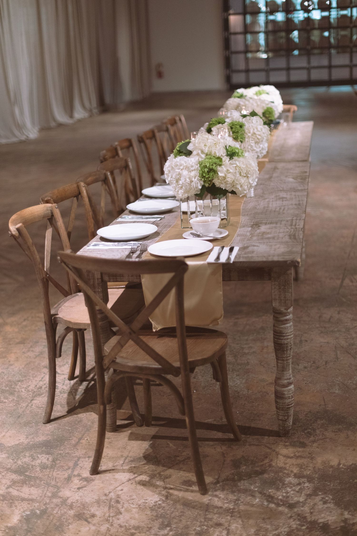 Rustic farmhouse wedding reception table with white hydrangea centerpieces and burlap runner on wood table.