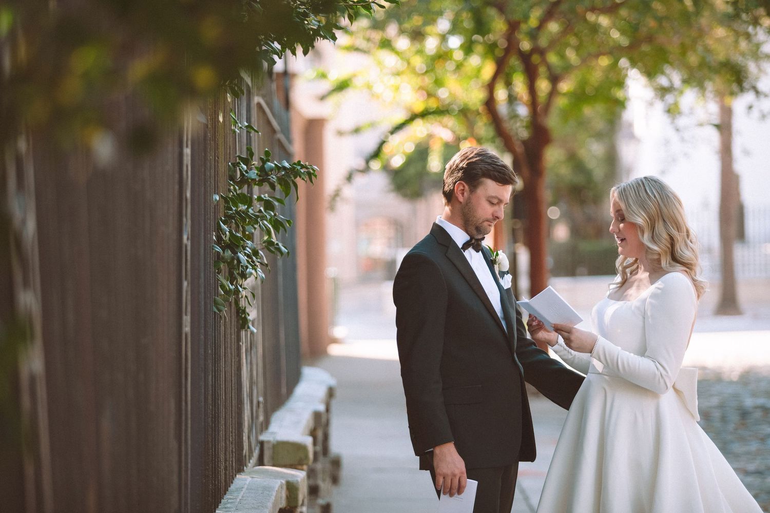 Bride in white gown reading vows to groom in black tuxedo along sunlit garden pathway.