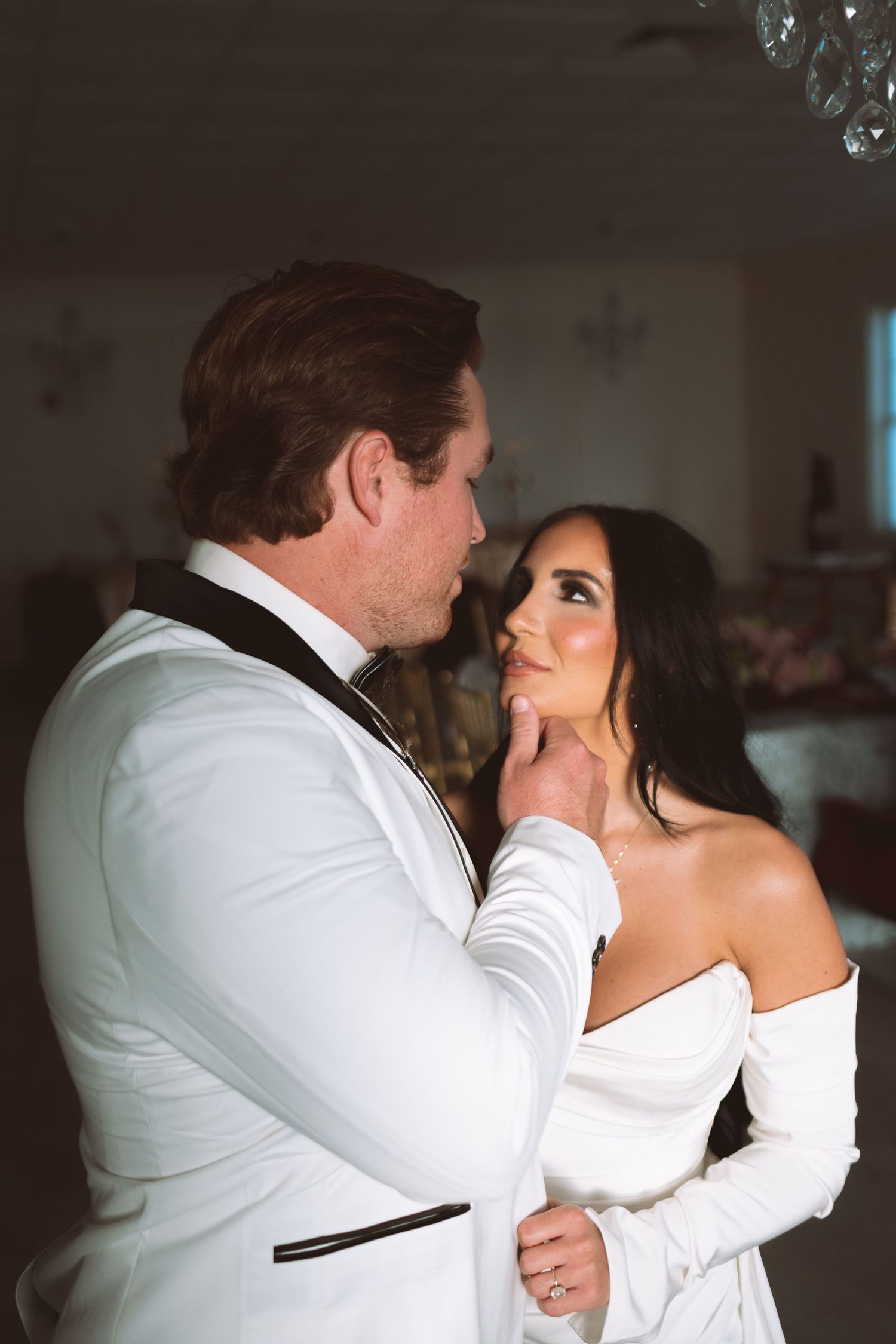 Groom in white tuxedo gently holds bride's chin during intimate first dance.
