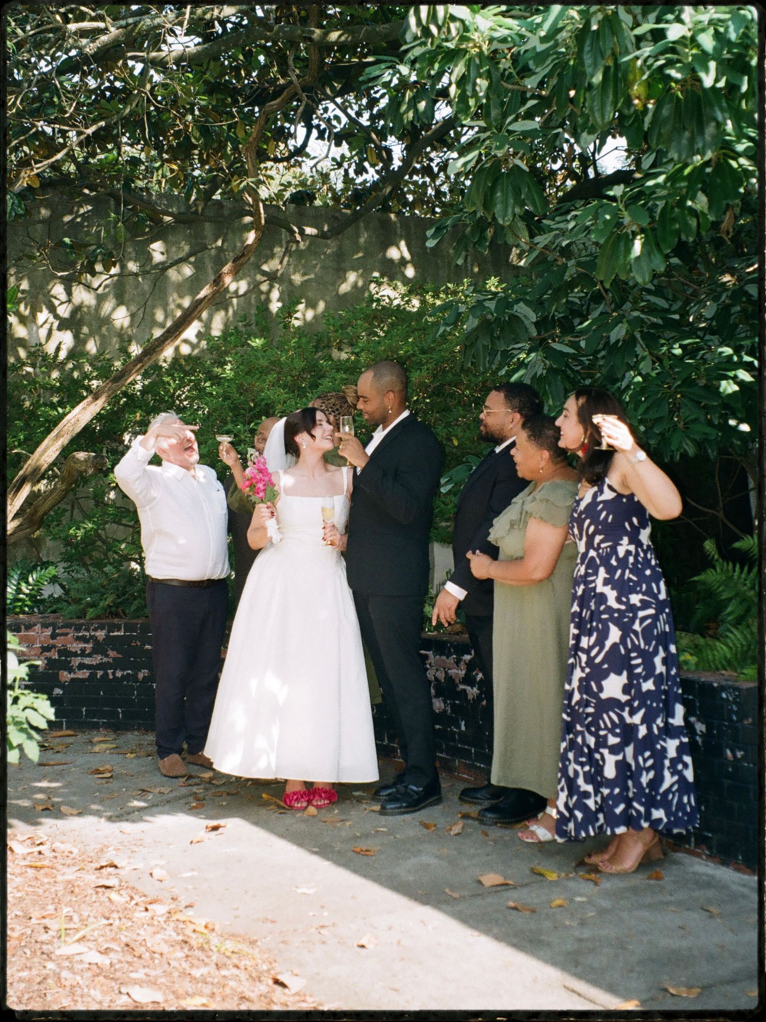 Wedding party celebrates with drinks beside brick wall under green trees in outdoor garden setting.