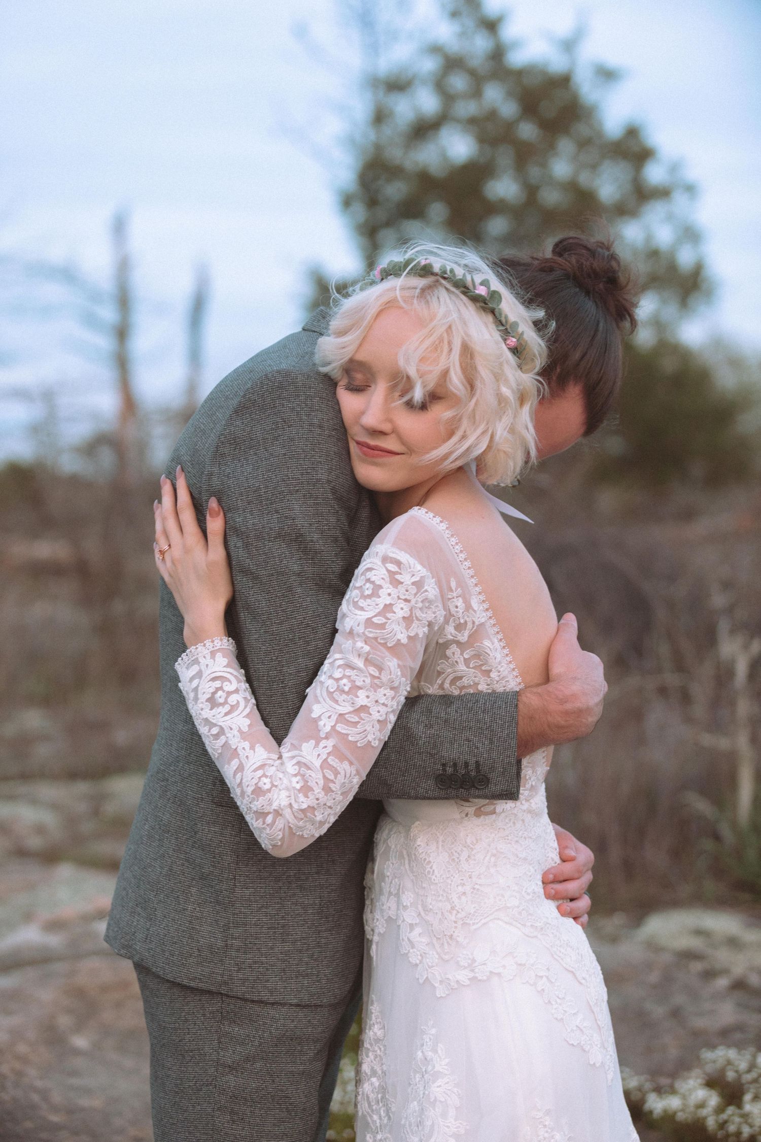 Bride in lace wedding gown embraces groom in gray suit outdoors during romantic wedding portrait.