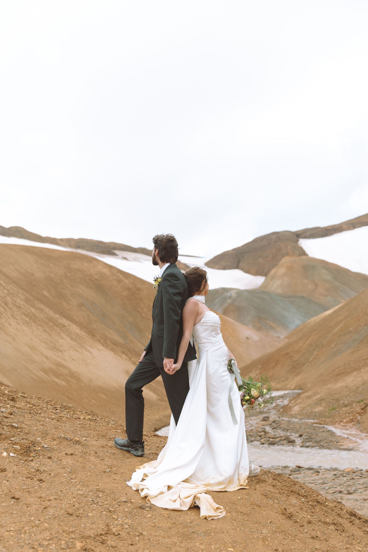 Newlyweds walk hand-in-hand along a rocky mountain path, bride in white gown holding a floral bouquet.