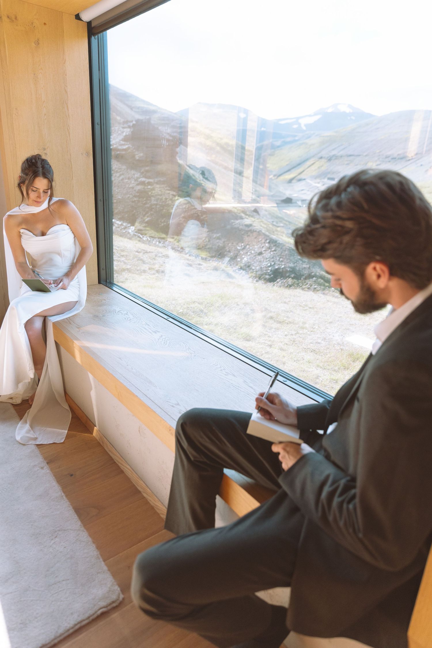 Bride and groom separately writing personal wedding vows by large window with mountain backdrop.