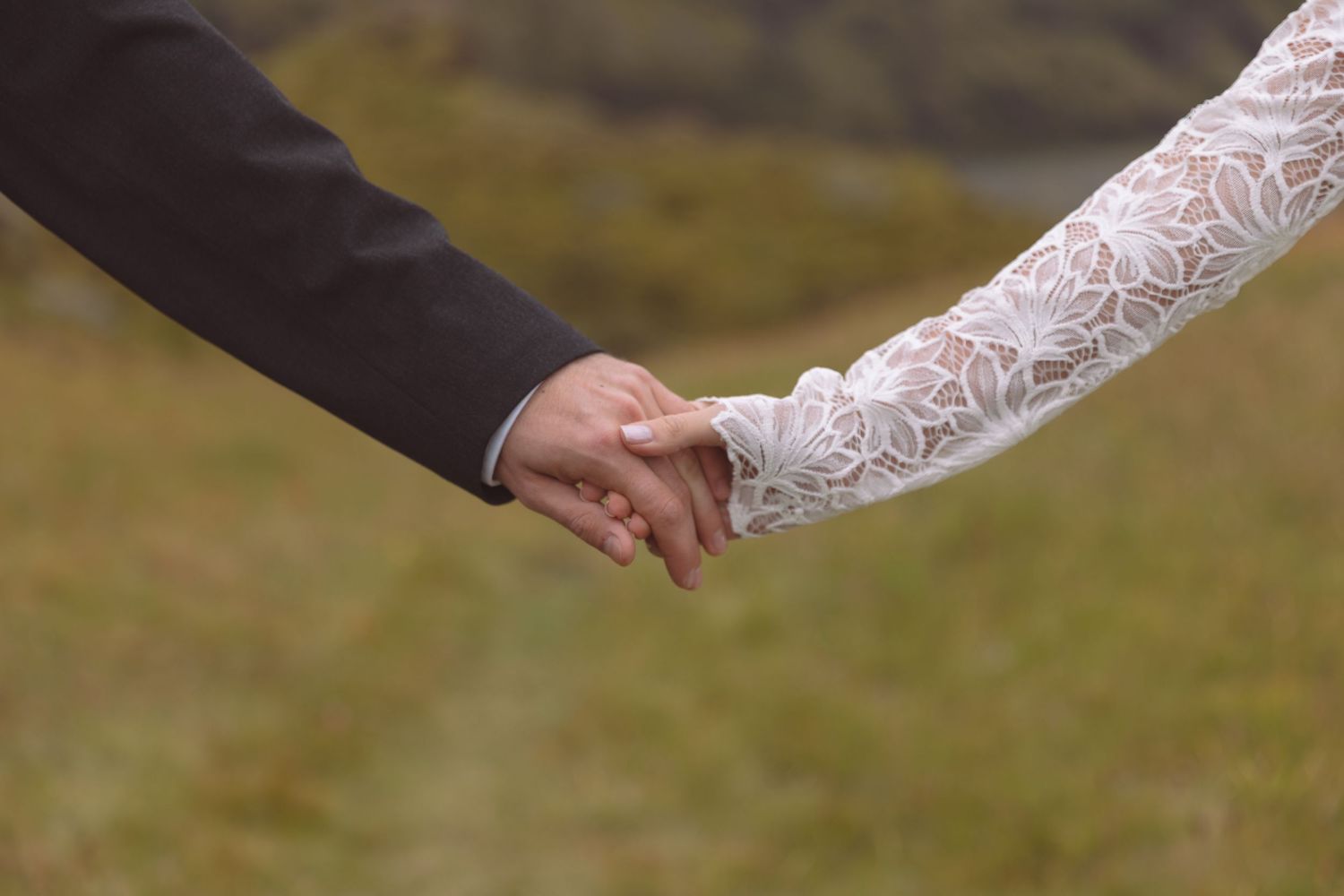 Close-up of bride and groom holding hands, lace wedding dress detail visible outdoors.
