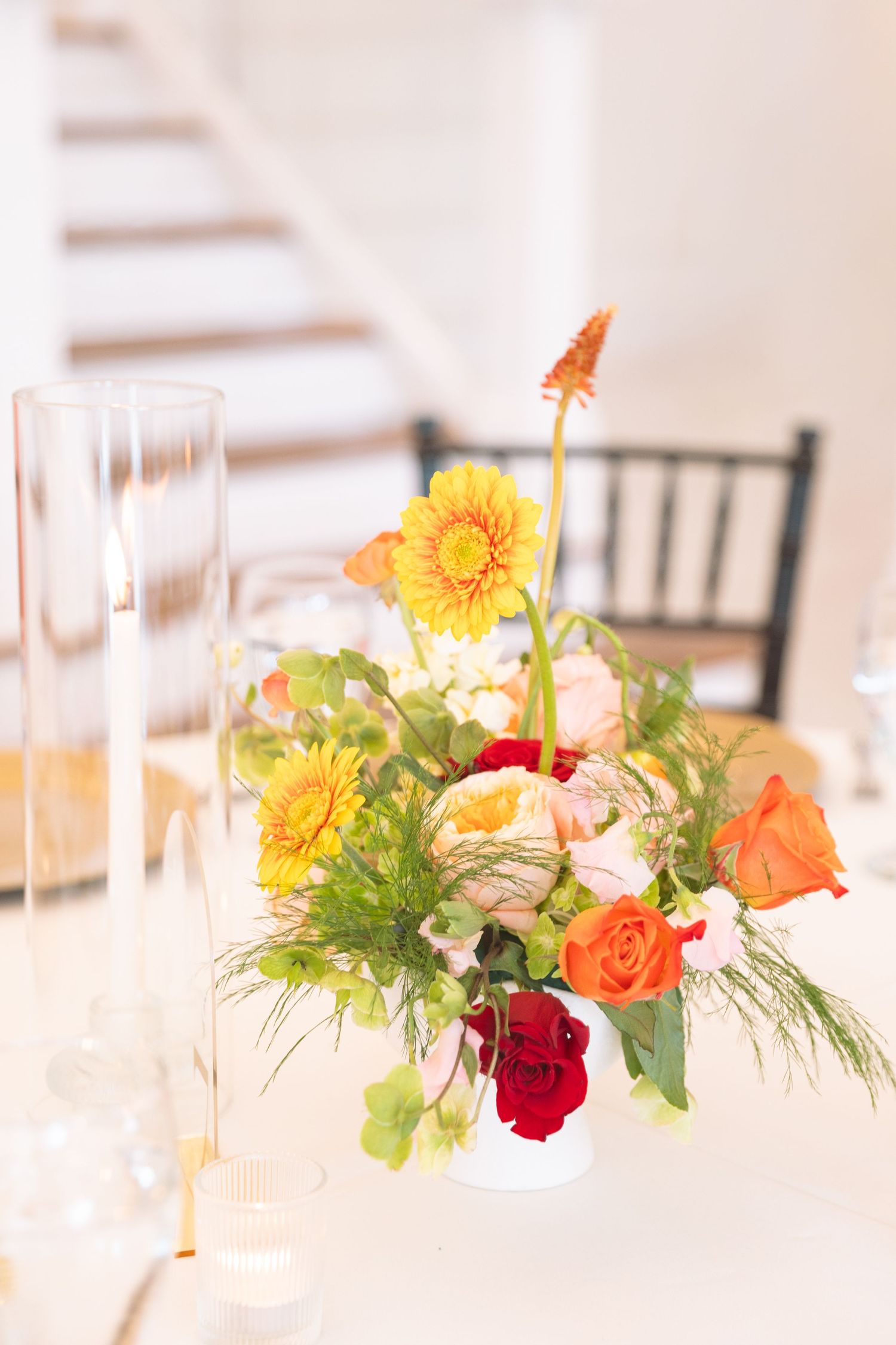 Vibrant centerpiece with yellow gerberas, orange and red roses in a white vase on a reception table.