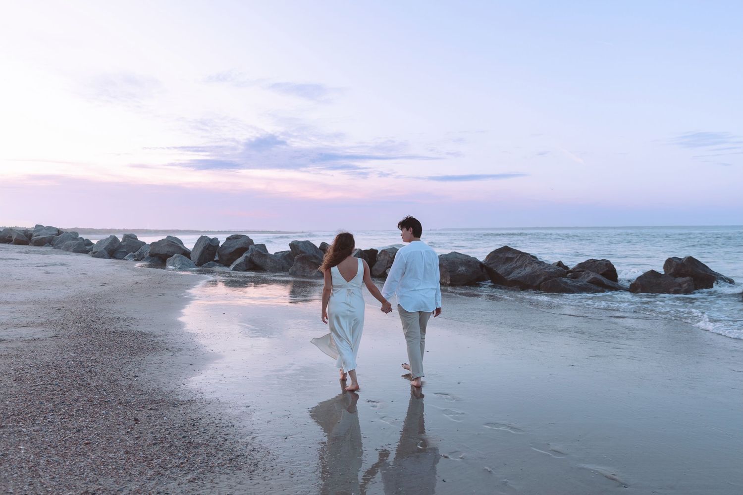 Couple holding hands walking along a reflective beach shoreline at dusk with rocky jetty nearby.