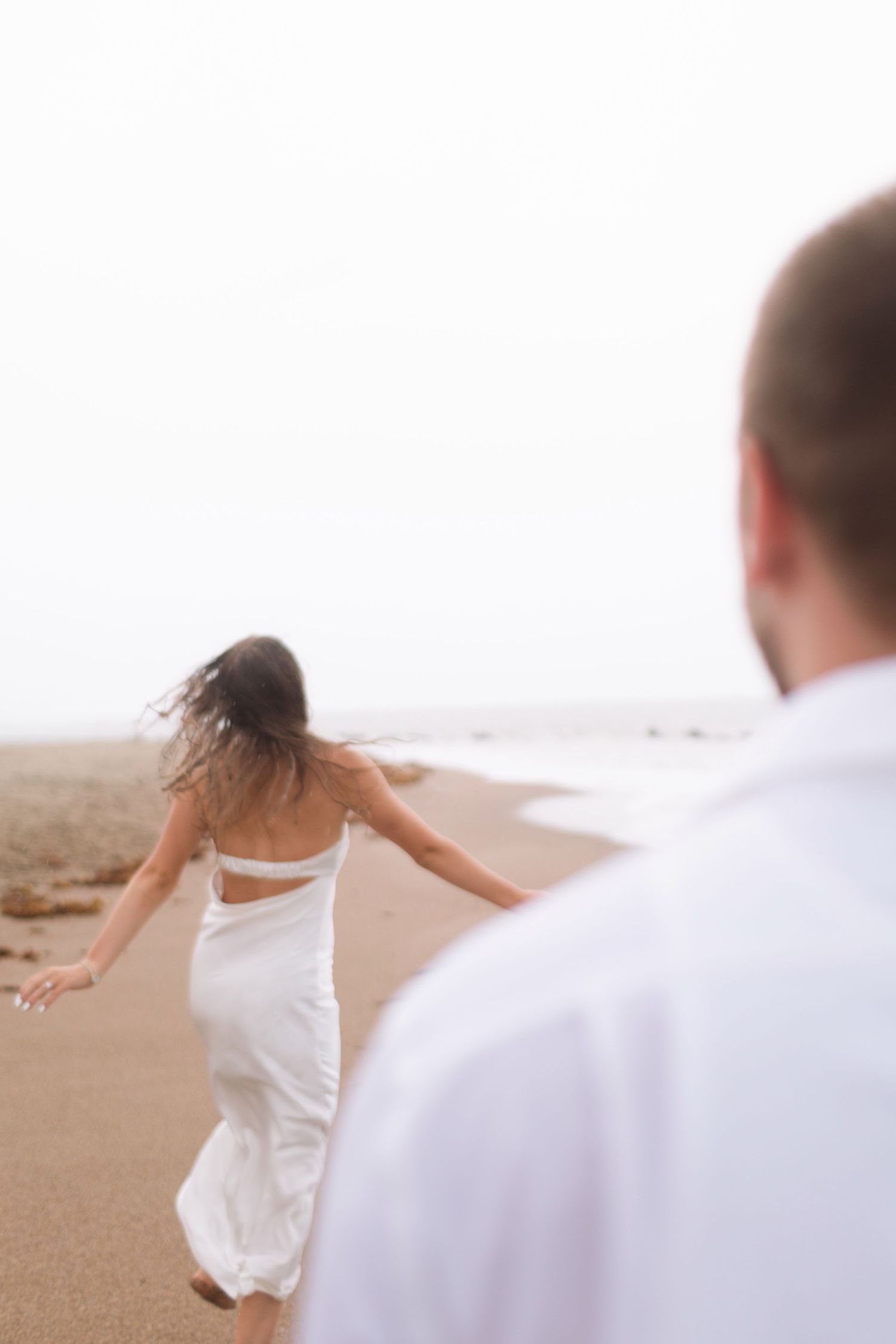 Woman in white dress running on beach while man watches from foreground.