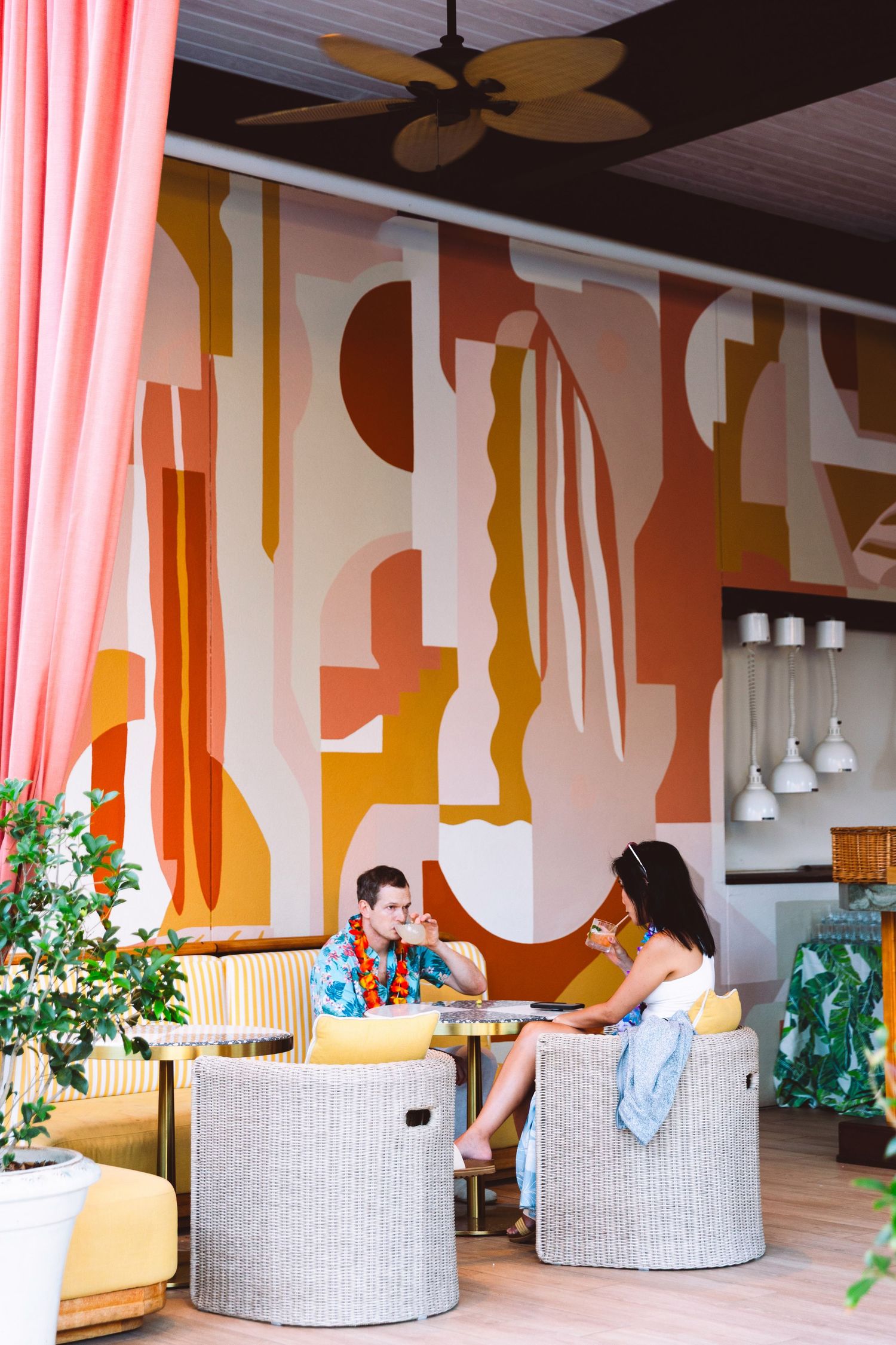 Couple relaxing in wicker chairs at a colorful tropical cafe with bold abstract mural and pink curtains.