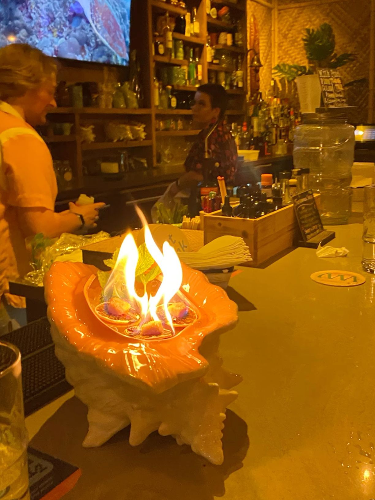 Flaming cocktail served in decorative shell at tropical tiki bar with bartender in background.