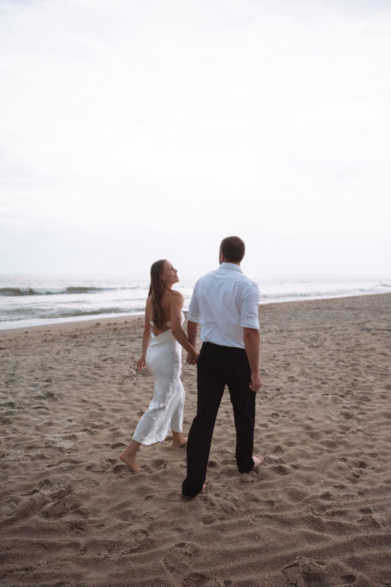 Couple holding hands walking barefoot on sandy beach toward ocean waves at dusk.