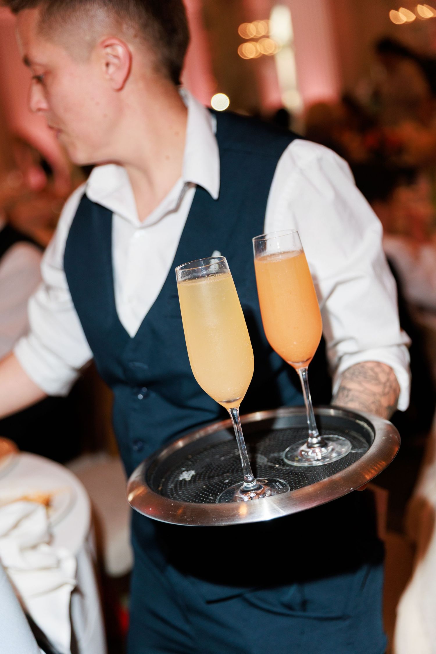 Waiter in vest carrying silver tray with two champagne flutes filled with orange mimosas at elegant event.
