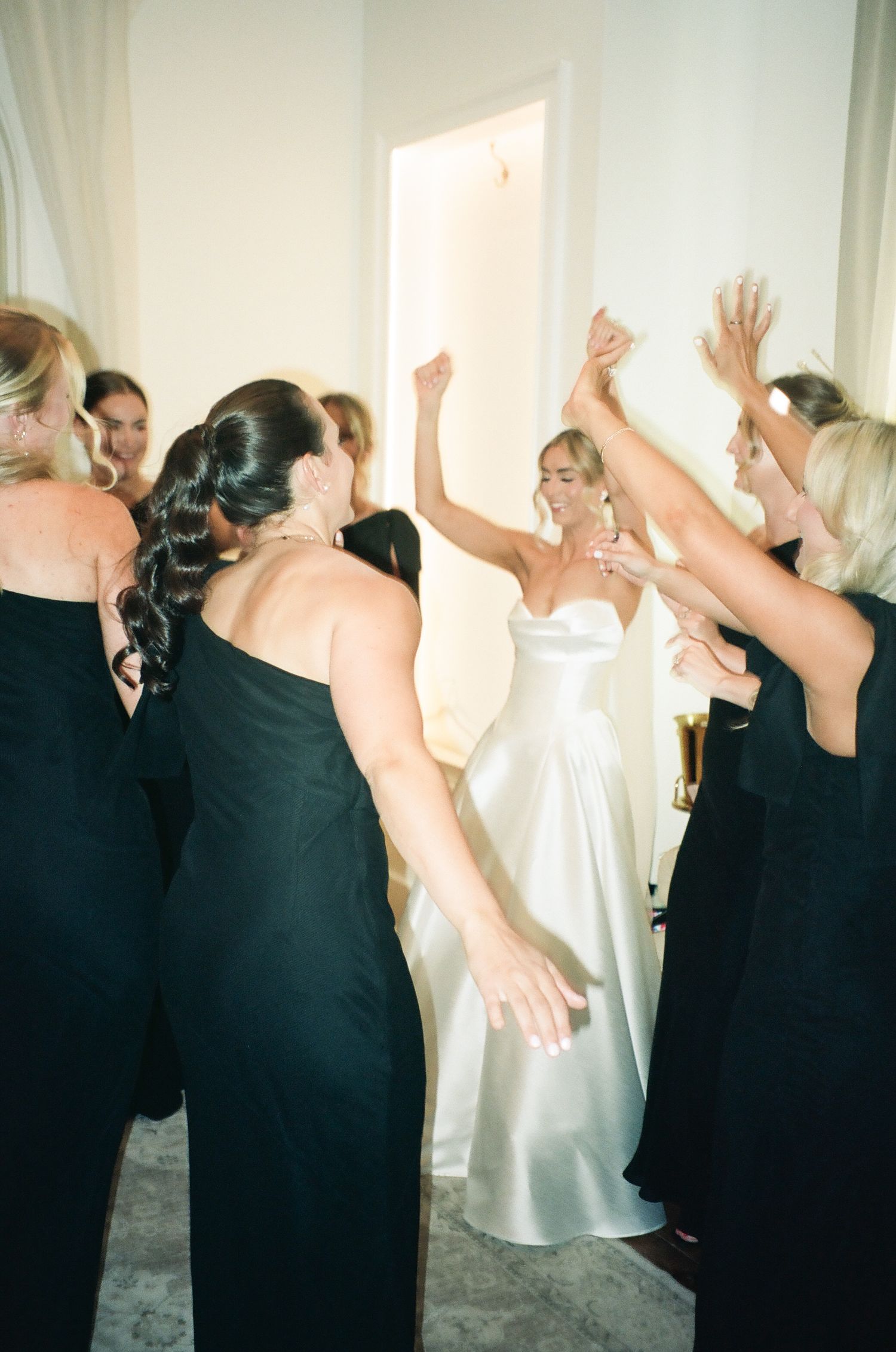 Bride in white satin gown celebrates joyfully with bridesmaids in black dresses, arms raised in excitement.