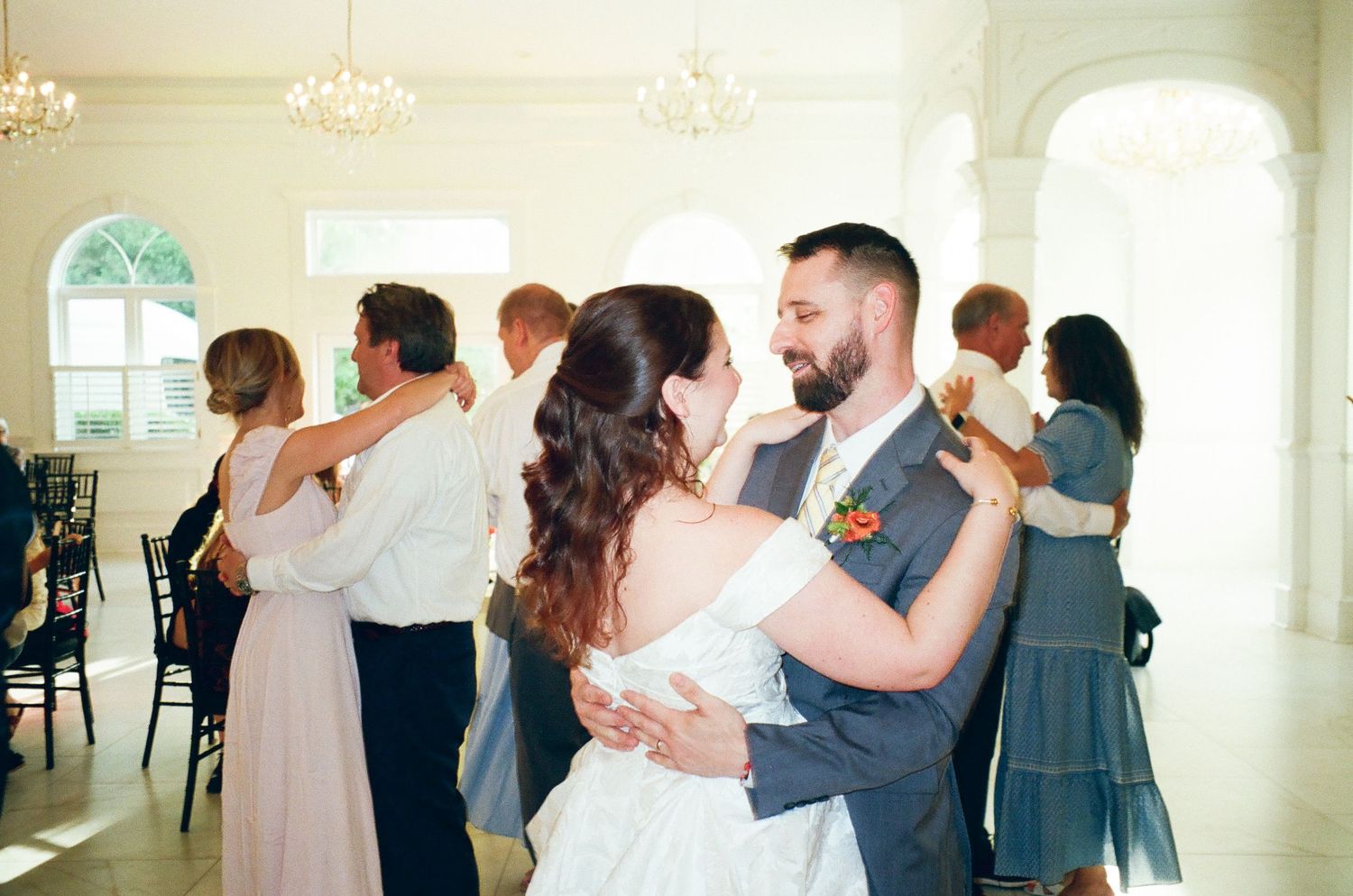 Bride and groom share first dance surrounded by guests in elegant white ballroom at wedding reception.