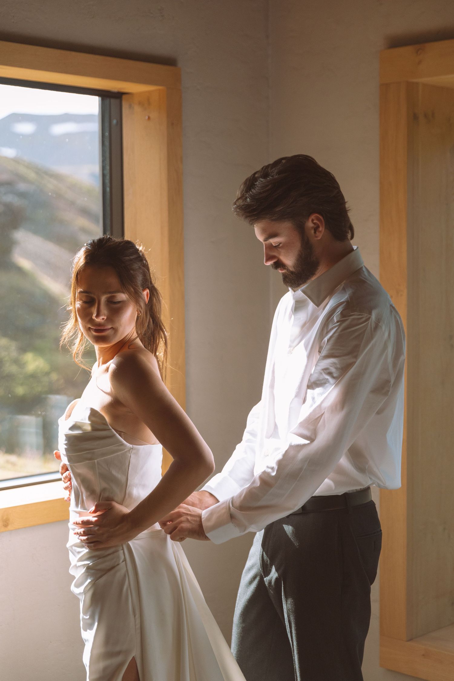 Groom adjusts brides white satin wedding dress while standing by sunlit mountain view window.