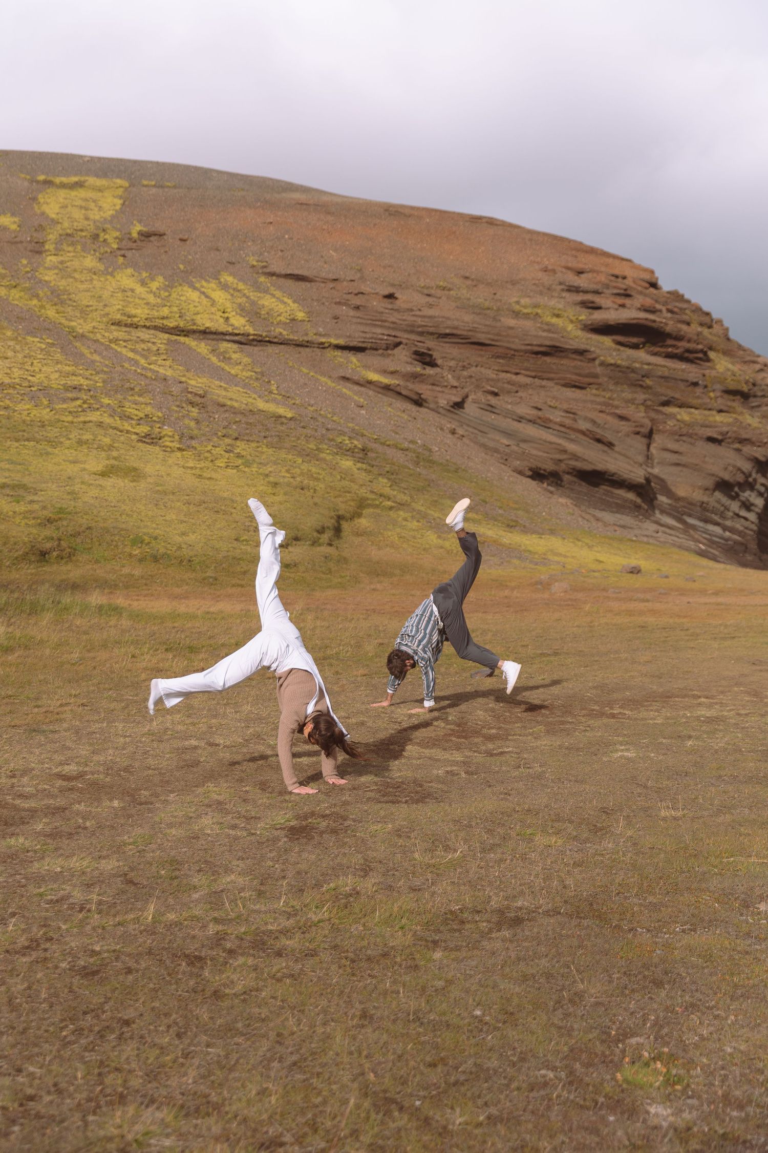 Two people doing cartwheels together on a green grassy field with rocky hills in the background.