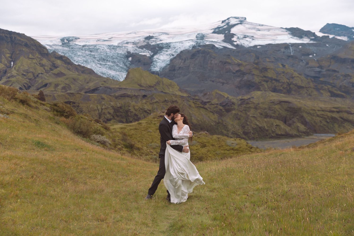 Bride and groom embracing romantically on grassy hillside with dramatic Icelandic glacier scenery.